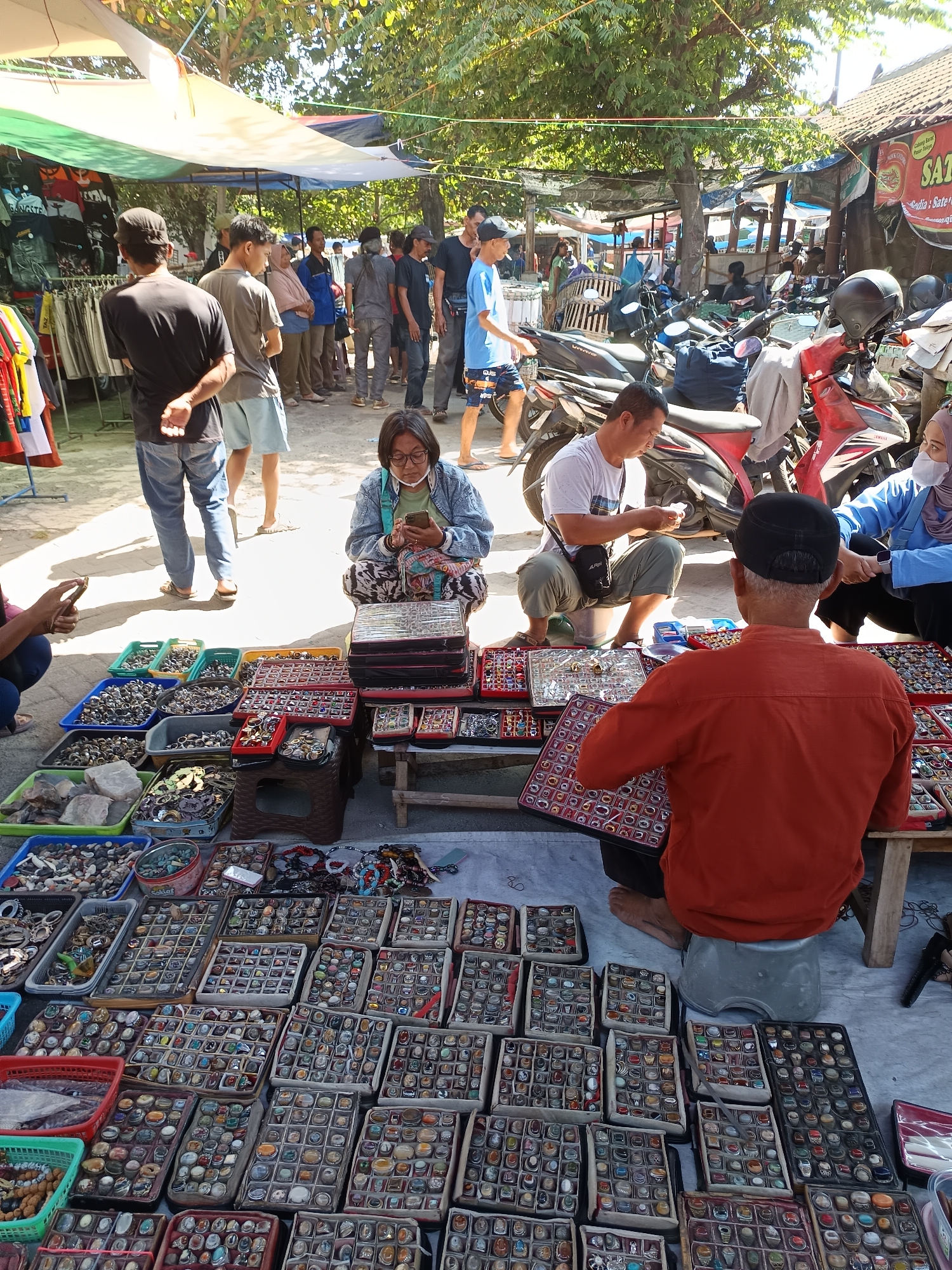 The image depicts an outdoor market scene in Geneng, Indonesia, likely a bustling local gathering place. The foreground is dominated by various stalls showcasing an array of goods, primarily rings displayed in numerous compartments within trays. These rings feature a variety of stone and metal designs. Several individuals are interacting with the merchandise or the vendors.
In the center, a woman wearing glasses and a denim jacket is seated, looking at her phone. To her right, a man in an orange shirt, likely a vendor, is examining a tray of rings. Behind him, a man in a white t-shirt is seated and appears to be engaged in a transaction. Further in the background, more people are browsing or walking through the market.
The market environment is characterized by makeshift stalls covered by tarps, providing shade from the sun. Several motorcycles are parked among the stalls, suggesting a mode of transportation for vendors and visitors. The ground is covered with a light-colored tarp or mat. The presence of trees overhead indicates an outdoor setting. The overall atmosphere appears to be lively and transactional, with vendors and customers engaged in commerce amidst a typical Indonesian market setting.