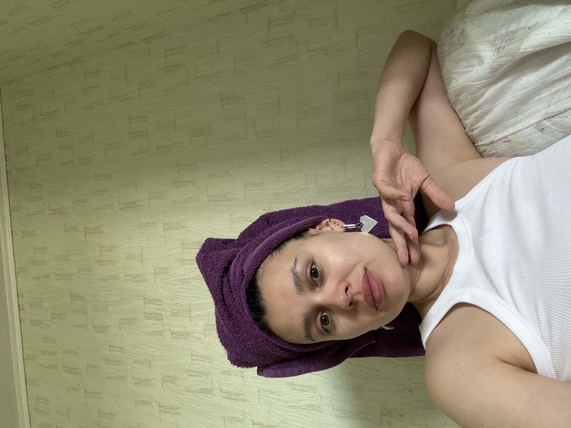 A person lies on a bed or similar soft surface with a white pillow. The person is wearing a white tank top and has a purple towel wrapped around their head. They are touching their neck with their fingers. The background consists of a light green wall with a subtle, repeating geometric pattern. The city of Chaikovsky, Russia is not discernible from the image.