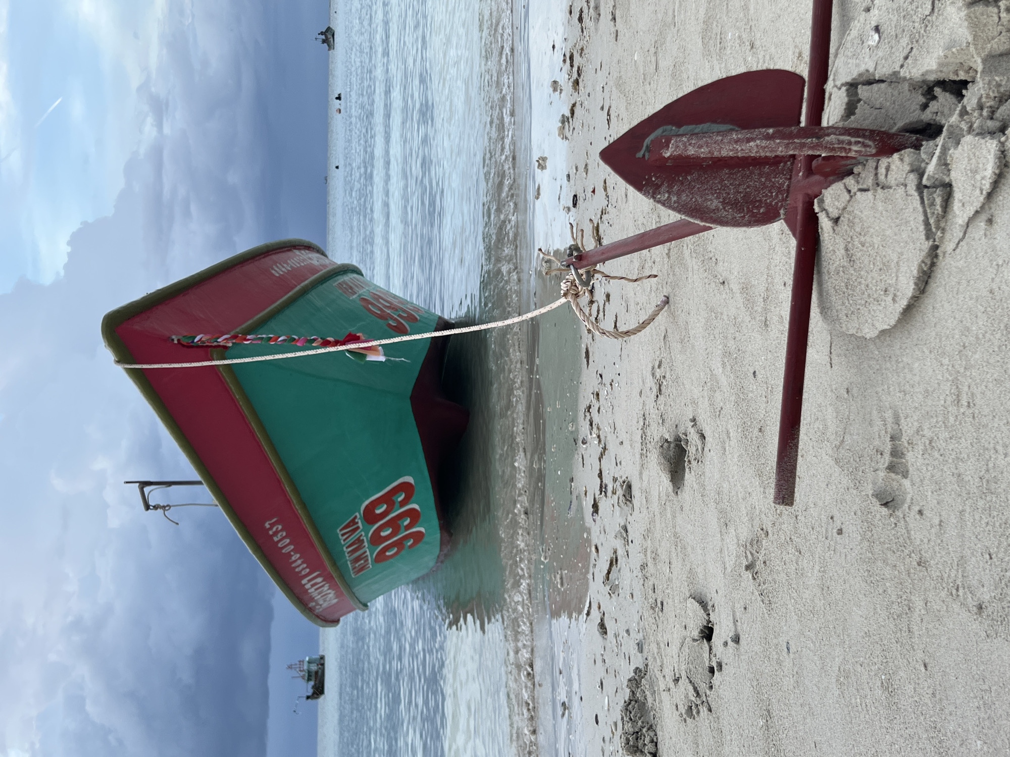 An anchor is partially buried in the sand on a beach in Ko Pha-ngan District, Thailand. A rope connects the anchor to a teal and red fishing boat that is partially submerged in the shallow water. The boat has the number "999" and the word "KENNAVA" painted on its side. In the distance, other boats are visible on the water. The sky is cloudy, with some patches of blue. The scene appears to be a calm, overcast day.