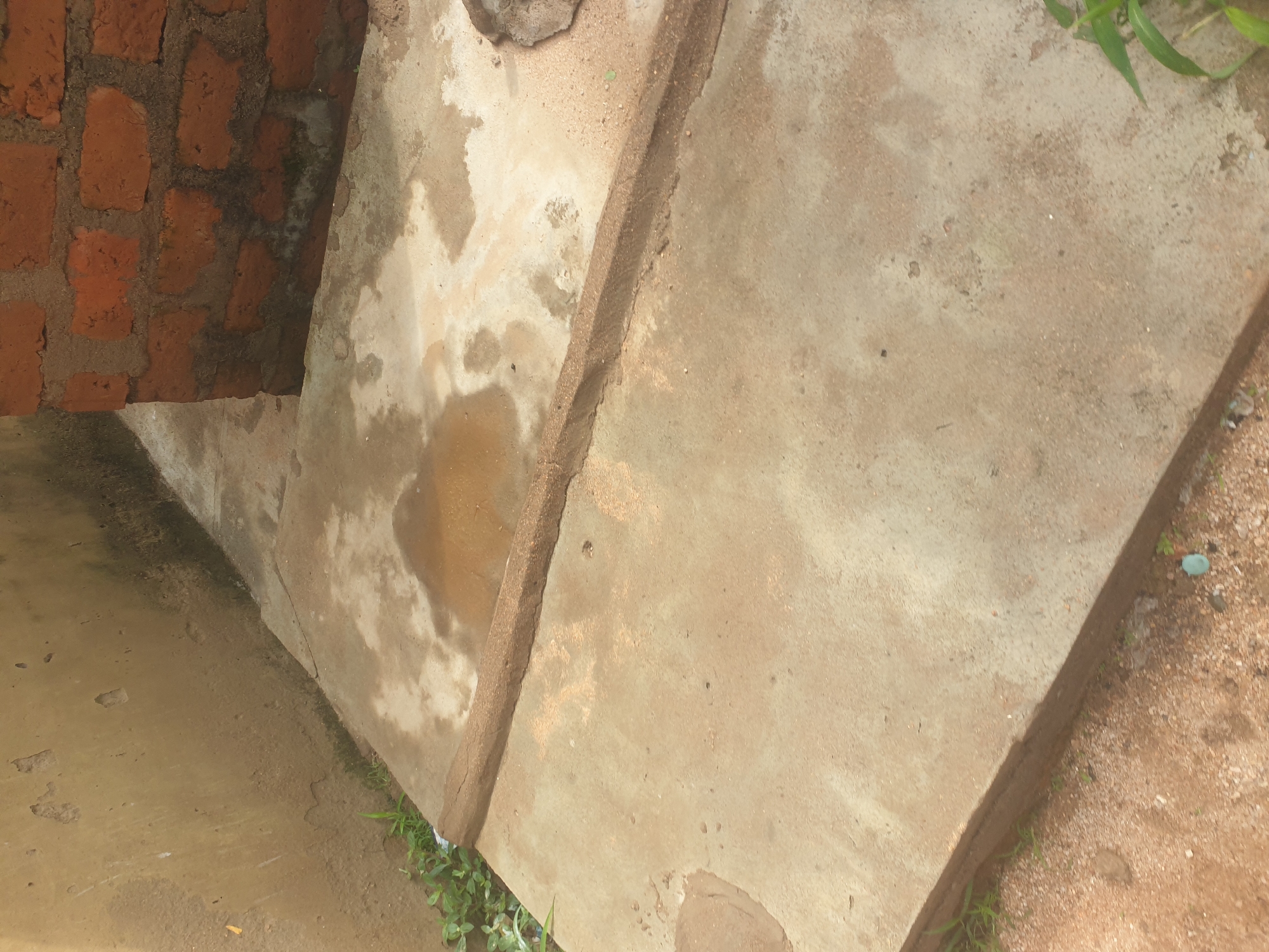 The image depicts an outdoor scene at ground level, likely in an urban or semi-urban area of Jalingo, Nigeria. The foreground features a rough, light brown concrete surface with a raised, darker brown concrete border running diagonally across it. This border has a textured appearance, suggesting it might be a drainage channel or a foundation edge.
To the left of the concrete surface, there is a beige-colored wall with some signs of wear and tear, including patches of missing material and darker streaks, possibly indicating moisture or algae growth. Some sparse green vegetation is visible at the base of this wall.
In the background, to the right, a section of a brick wall constructed from reddish-brown bricks is prominent. This brick wall also shows some dark discoloration near its base. The ground continues to be a mix of concrete and what appears to be sandy soil. There are water stains or damp patches on the concrete surfaces. The overall impression is one of an unrefined, utilitarian outdoor space, possibly part of a building's exterior or a yard. No people or animals are visible in the image.