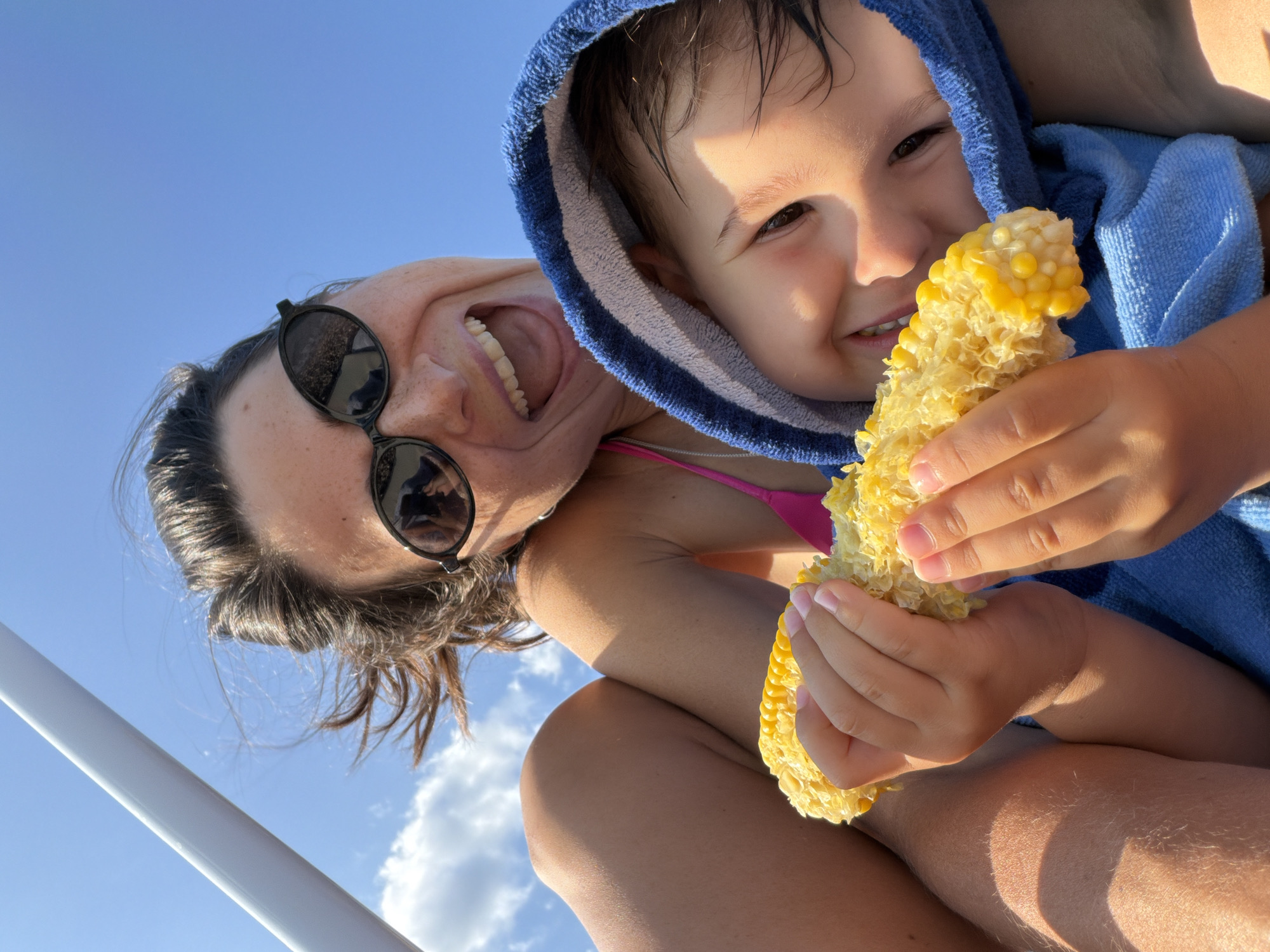In Budva, Montenegro, a low-angle, eye-level shot captures a woman and a child outdoors against a bright blue sky with scattered clouds. The woman, wearing black sunglasses and a pink bikini top, is looking up and smiling broadly. Her dark hair is slightly windswept. She is holding a partially eaten ear of corn. A child, wrapped in a blue hooded towel, is positioned in front of the woman, also smiling and holding the corn. The child's face is illuminated by sunlight, and their dark hair appears wet. The scene suggests a casual, possibly beachside, outing.