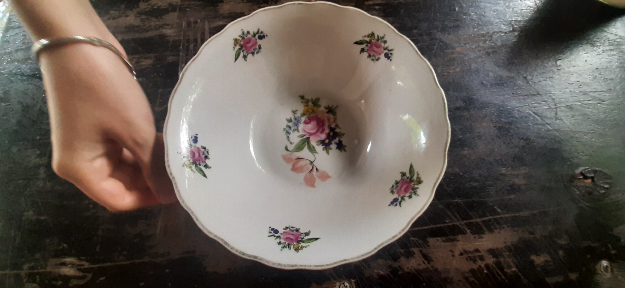 An overhead shot shows a white, scalloped-edge bowl with a floral pattern, resting on a dark, weathered wooden surface. A person's left hand, adorned with a thin, metallic bracelet, is visible on the left side of the frame, partially obscuring the bowl. The bowl itself is decorated with clusters of pink, purple, and yellow flowers with green leaves. The wooden surface appears to be a table, with visible grain and some scratches. The overall scene is a still life, focusing on the bowl and the hand. This image was captured in Yessentukskaya, Russia.