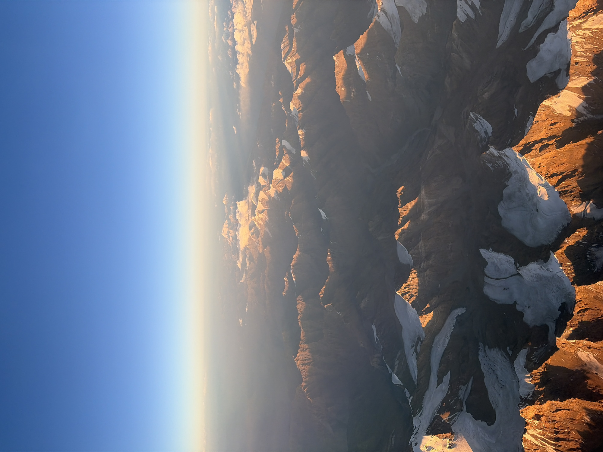 An aerial view captures a vast mountain range under a clear blue sky transitioning into a hazy horizon. The mountains, characterized by rugged, shadowed peaks and snow-capped summits, are illuminated by the warm, golden light of what appears to be sunrise or sunset. The varying angles of light and shadow accentuate the dramatic topography, revealing deep valleys and sharp ridges. A layer of haze softens the distant peaks, creating a sense of depth and scale. The scene is devoid of any signs of human activity or structures, presenting a natural, unpopulated landscape. The location is identified as Unknown, Tajikistan.