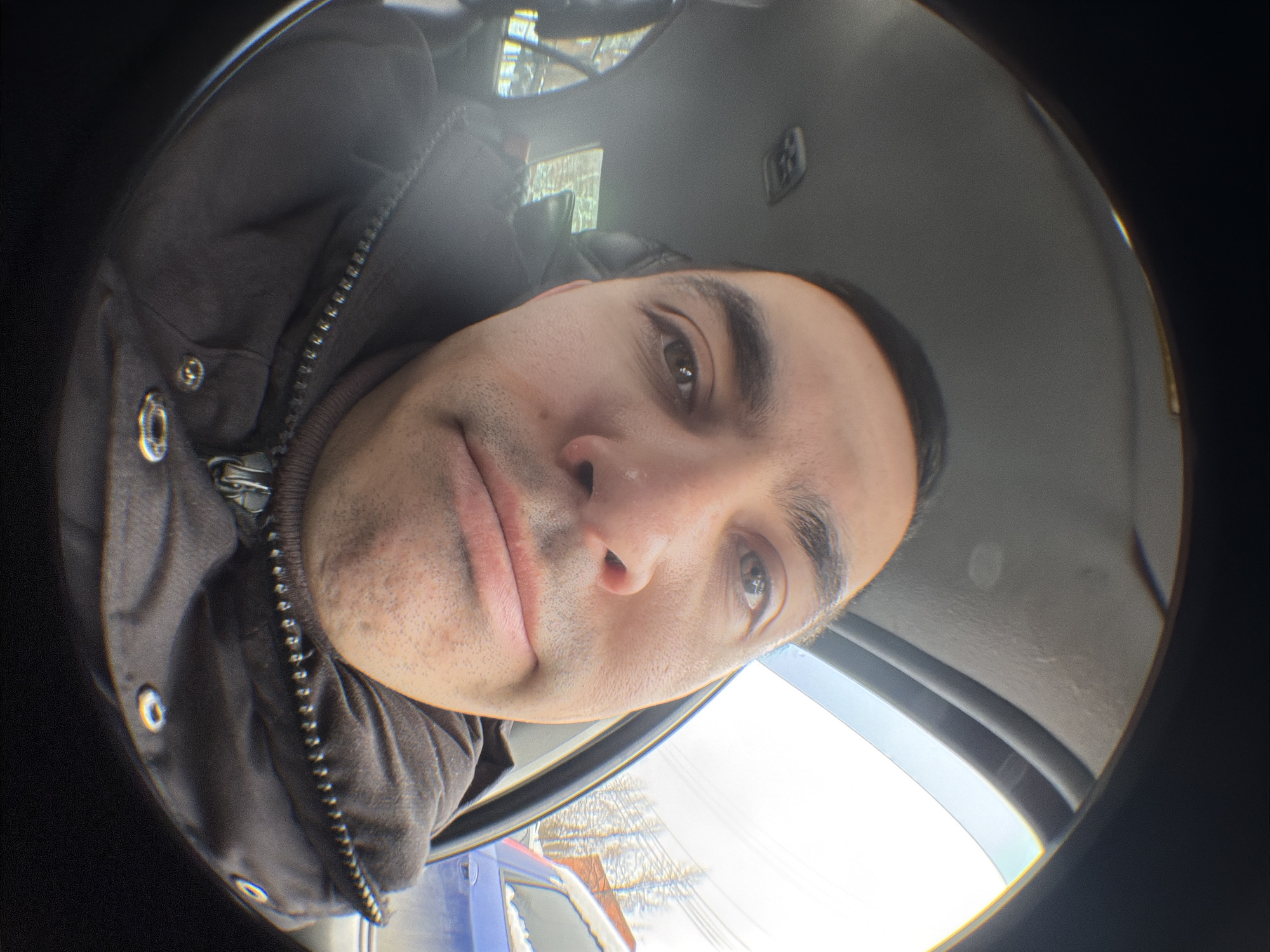 The image shows a close-up selfie of a man in what appears to be a vehicle. The man has dark hair, brown eyes, and light skin. He is wearing a dark jacket with a visible zipper and metal buttons. The photo has a distorted, fisheye lens effect. The background visible through the vehicle's windows shows a street scene, possibly in winter, with buildings, trees, and a blue car partially visible. Based on the information provided, the location is presumed to be Izhevsk, Russia. The image captures the man's face and upper body inside the vehicle, with the surrounding environment visible through the windows.