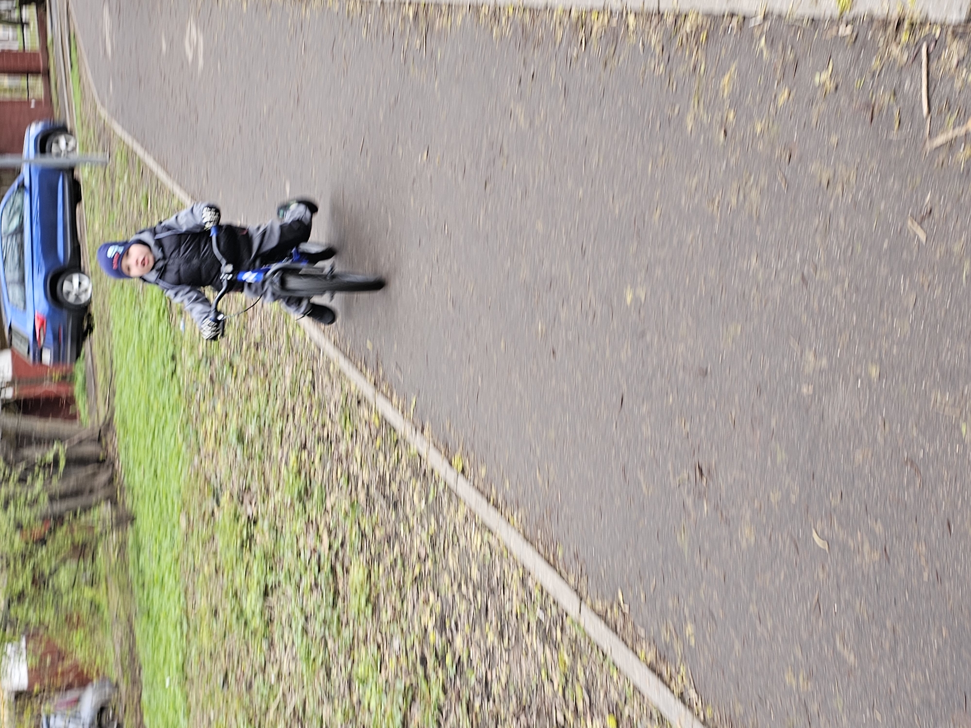 A young child is riding a small bicycle on a paved pathway. The child is wearing a dark coat, grey pants, gloves and a blue hat. The pathway runs alongside a green area with trees, and fallen leaves are scattered on the ground. A blue car is parked nearby. The scene is in Moscow, Russia.