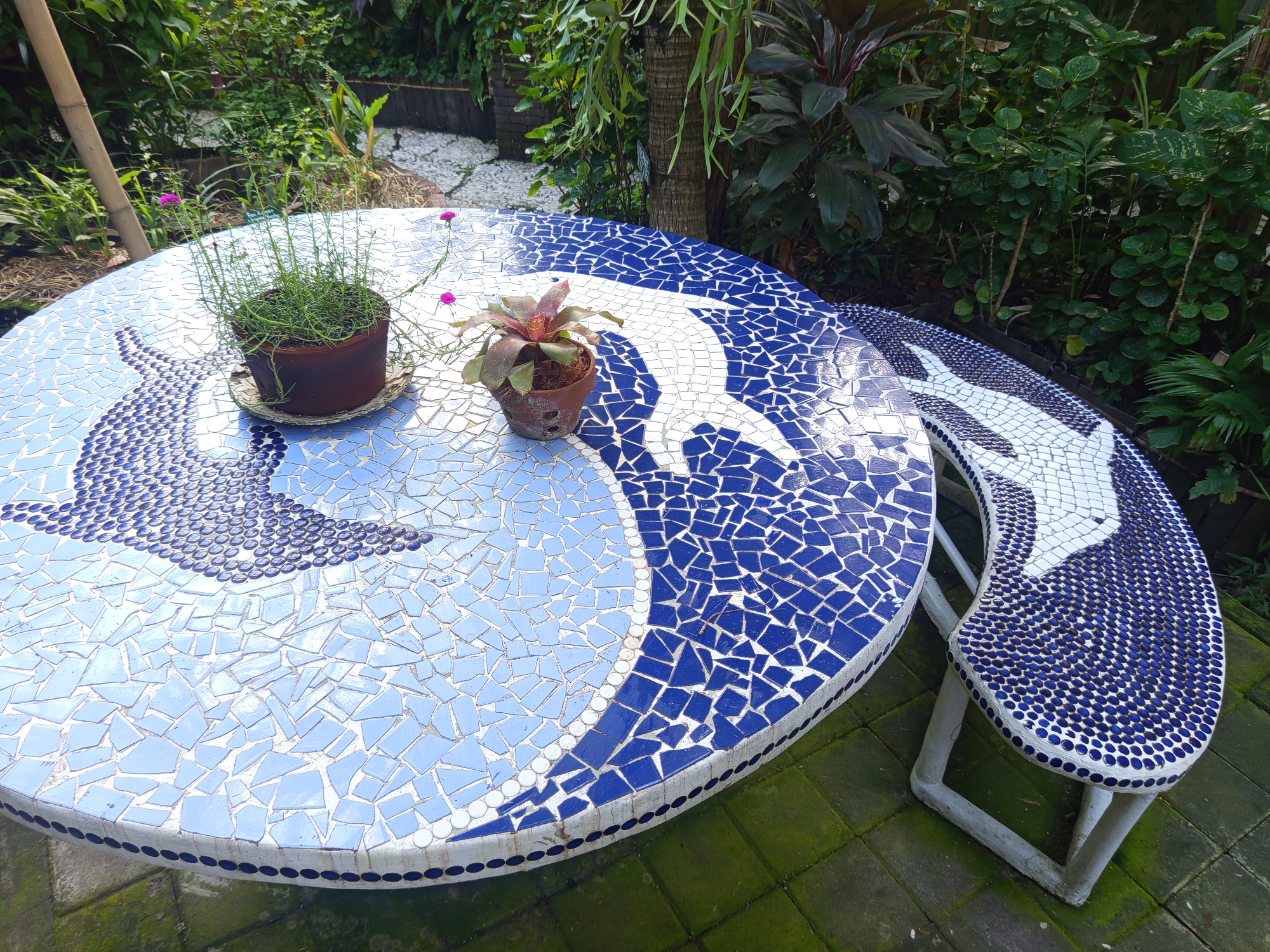 The image shows a round mosaic table with a matching curved bench in a garden setting in North Kuta, Indonesia. The table and bench feature a dolphin motif in white and blue tiles. Two potted plants sit on the table. Lush greenery surrounds the table and bench. The paving under the table and bench is green and mossy.