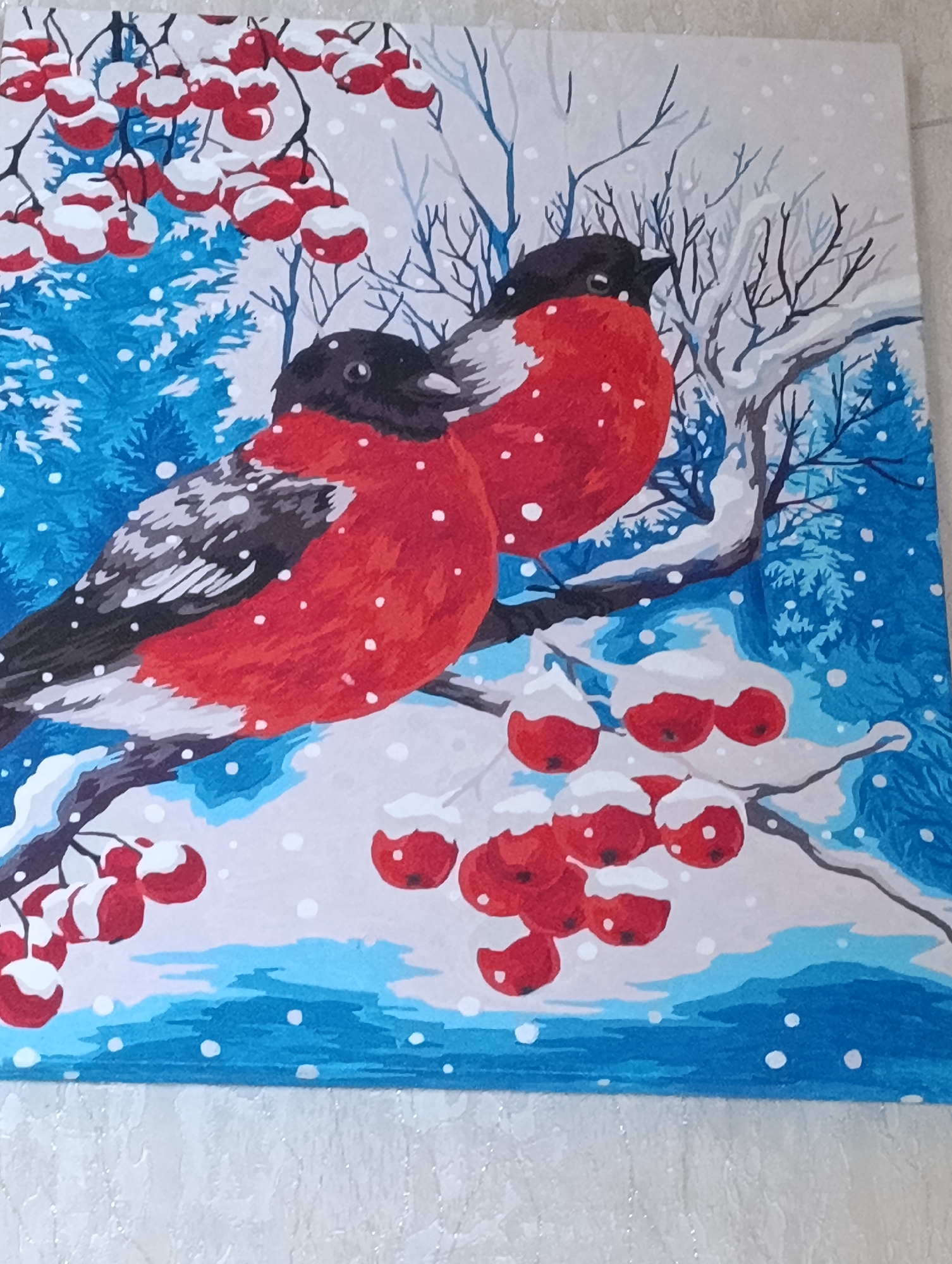 A painting depicts two bullfinches perched on a snow-covered branch with clusters of red berries. The background features a snowy forest scene with blue pine trees and bare branches. Snowflakes are falling throughout the image, creating a winter atmosphere. The painting is rendered in a stylized, illustrative manner with bold colors and defined outlines.