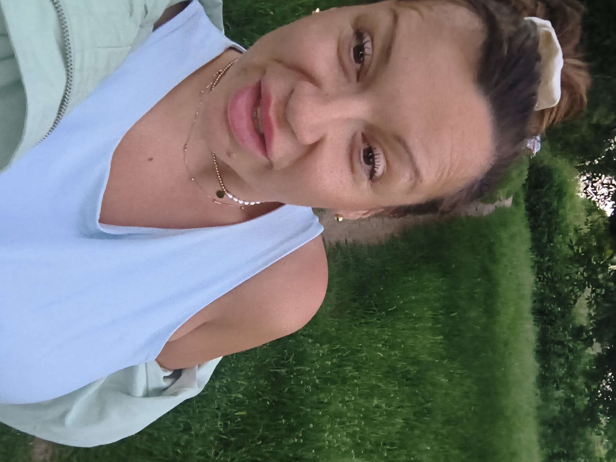 A woman is standing in a grassy area, possibly a park or yard. She is wearing a light blue sleeveless top and a light green jacket. She has dark hair pulled back with a light-colored scrunchie or hair tie. She is wearing a delicate gold necklace and small gold earrings. The background consists of green grass and foliage, suggesting an outdoor setting. The location, Sofia, Bulgaria, is not confirmed visually but has been included.