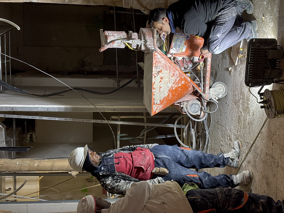 The scene depicts an active, dimly lit indoor construction site in Madrid, Spain. Three adult males are present, illuminated primarily by an LED floodlight on the concrete floor in the foreground, connected to a yellow cable reel.
On the left, a man with a white or light-colored cap and light-colored jacket is partially visible, facing towards the center. The central figure, wearing a white hard hat, a dark patterned hoodie over a red garment, and dark blue work pants with orange pocket accents, stands with his hands in his pockets. A small tool is clipped to his right hip. His clothing and work boots are soiled with dust and debris. He appears to be observing the area to his right or the man crouching.
To the right, a third man is crouched beside an orange mobile construction machine, likely a plastering or mortar mixing unit. This machine features a large, dirtied hopper filled with a grey powdery substance, wheels, a motor, and various connected hoses and electrical cables. The man is wearing a dark jacket and dark pants, with a dark blue scarf around his neck. He has a small, dark object held between his lips while he inspects the machine's lower section.
The background shows unfinished walls with exposed metal stud framing, visible pipes, electrical conduits, and general construction debris. Portions of bags, possibly containing construction materials, are visible on the far right. The overall environment is dusty and suggests ongoing renovation or building work.