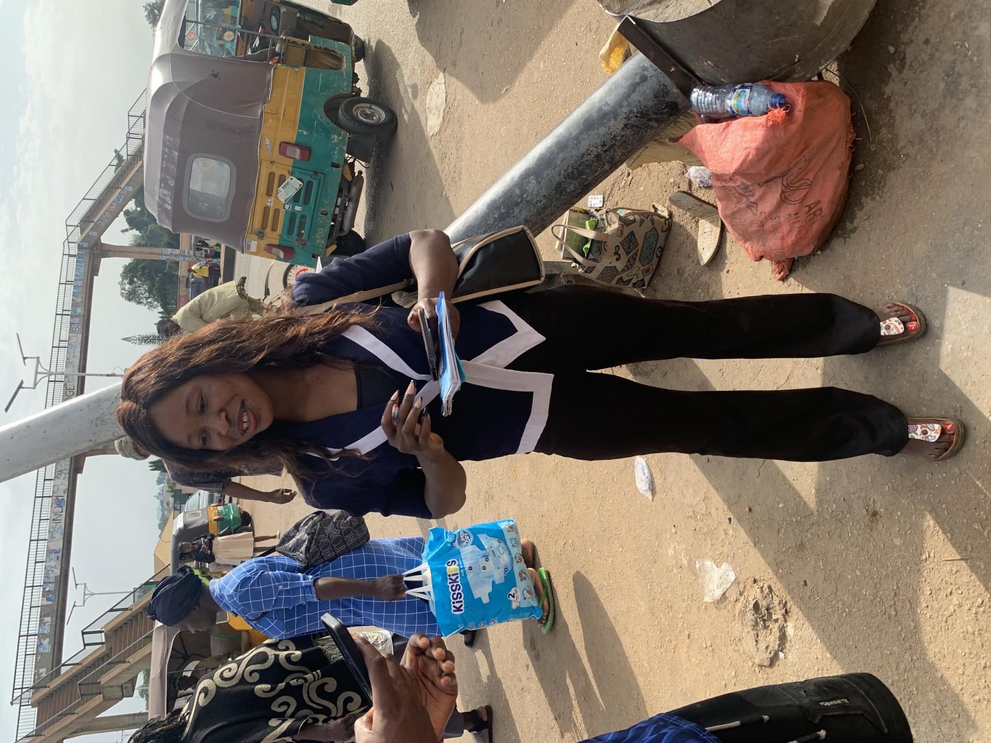 The image shows a street scene in Gura Top, Nigeria. A woman in a dark blue jacket with white trim and black pants is standing in the foreground, holding a small stack of papers and a pen. She is looking to her right and smiling. Behind her, other people and vehicles are visible. To her left, a person in a blue and white checkered shirt is holding a blue shopping bag with "Kisski's" written on it. A green and yellow auto-rickshaw is parked behind them. In the background, a pedestrian bridge crosses over the street. The ground is paved with dirt and asphalt, and there are some pieces of trash scattered around.