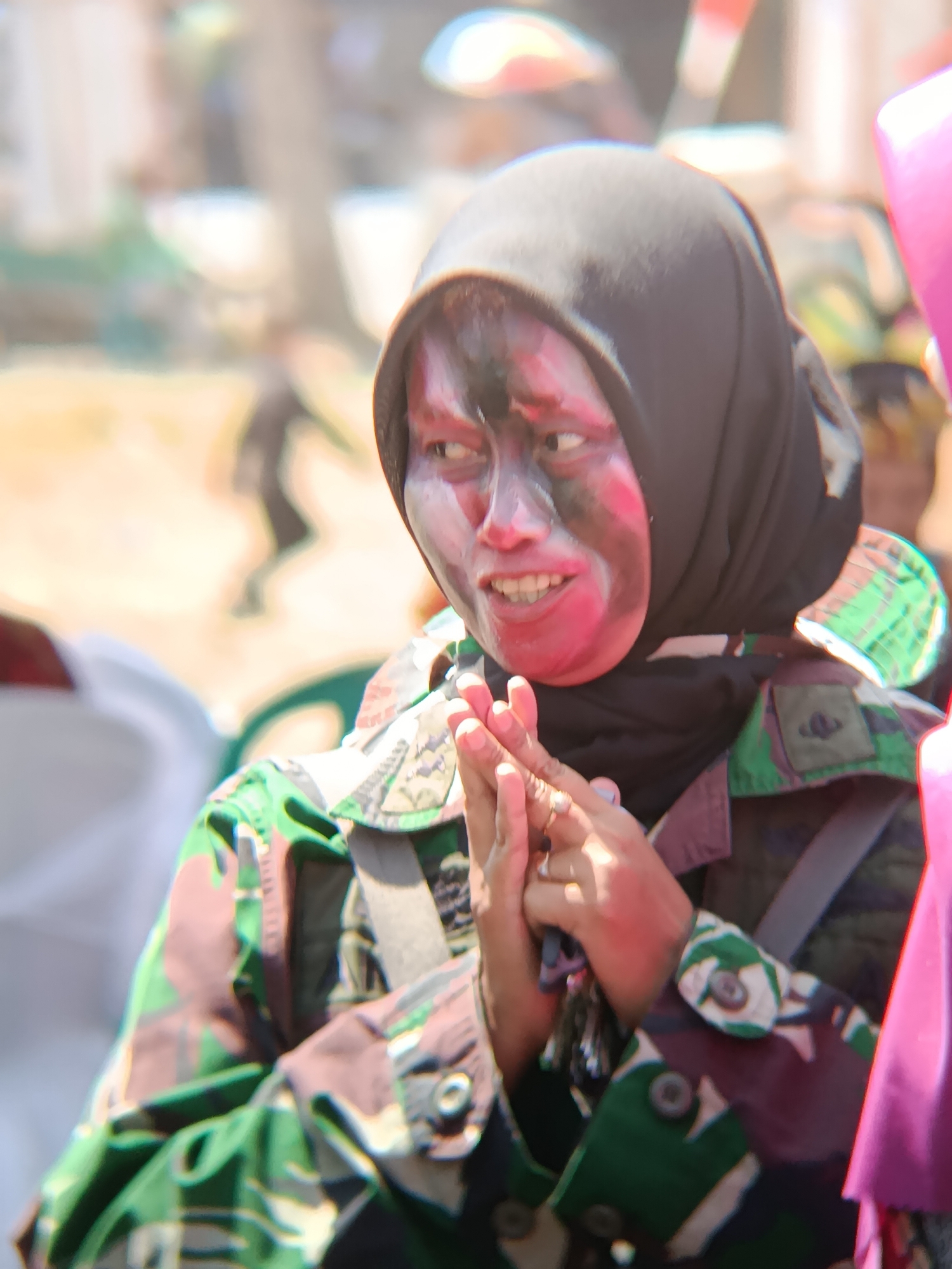 A person wearing a black hijab and camouflage uniform with face paint in a desert or dirt-like setting, possibly during an event or demonstration. The person's hands are clasped together in front of their chest. The background is blurred with hints of other people and structures. The lighting suggests it is daytime.
