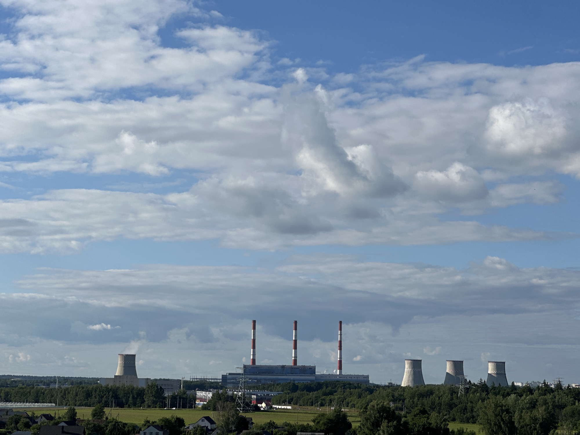 This is a horizontal, eye-level, medium-wide shot of a power plant on a sunny day, with a backdrop of a cloudy blue sky. The power plant, located in the Шчомысліцкі сельскі Савет, Belarus, features several structures, including three tall, red-and-white striped chimneys, a large, long, grey building, and three large, concrete cooling towers. One of the cooling towers on the far left is emitting a plume of white steam. The power plant is surrounded by a dense forest, with some residential buildings visible in the foreground. The sky is filled with a mix of white, fluffy clouds and patches of bright blue. The sun appears to be high in the sky, suggesting it is daytime. The overall impression is one of industrial power within a natural landscape. There is no visible text in the image.