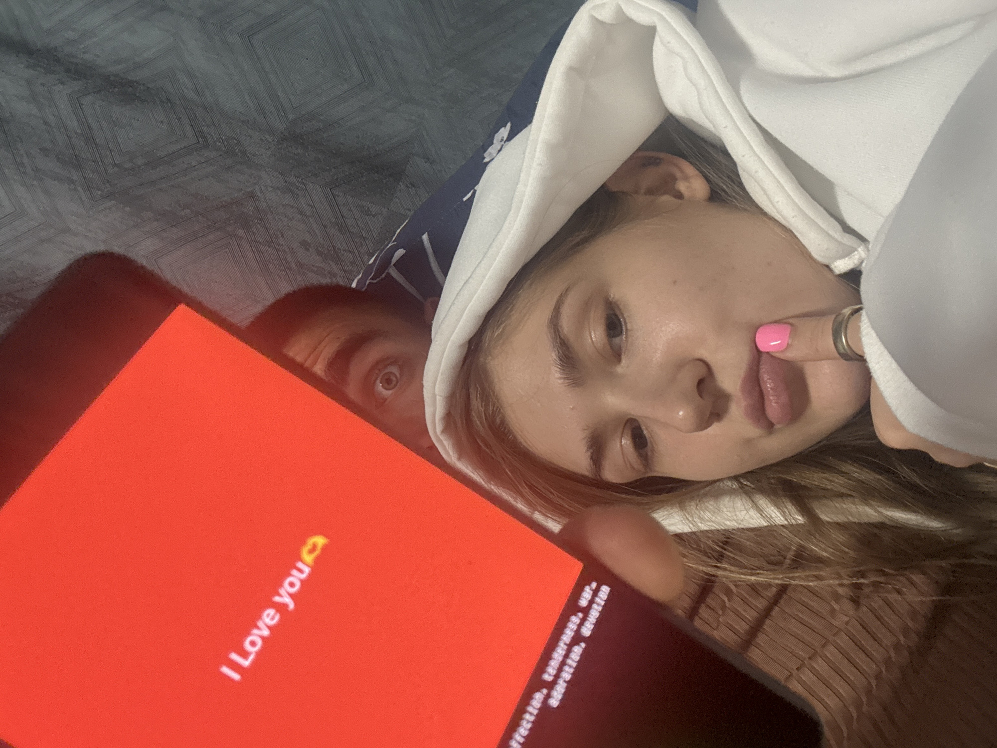 The image is a selfie taken indoors, possibly in a bedroom, with a predominantly red screen from a phone in the foreground displaying "I Love you" with a kissing emoji. A young woman with blonde hair, wearing a white hoodie, is looking at the camera, with her finger touching her chin and a bright pink nail visible. A man's eye and forehead are visible behind her, peeking from under a patterned blue and white blanket. The background includes a textured grey surface and a woven brown surface, likely furniture or bedding. The image does not provide any specific information about the location in the city of Ussurijsk, Russia.