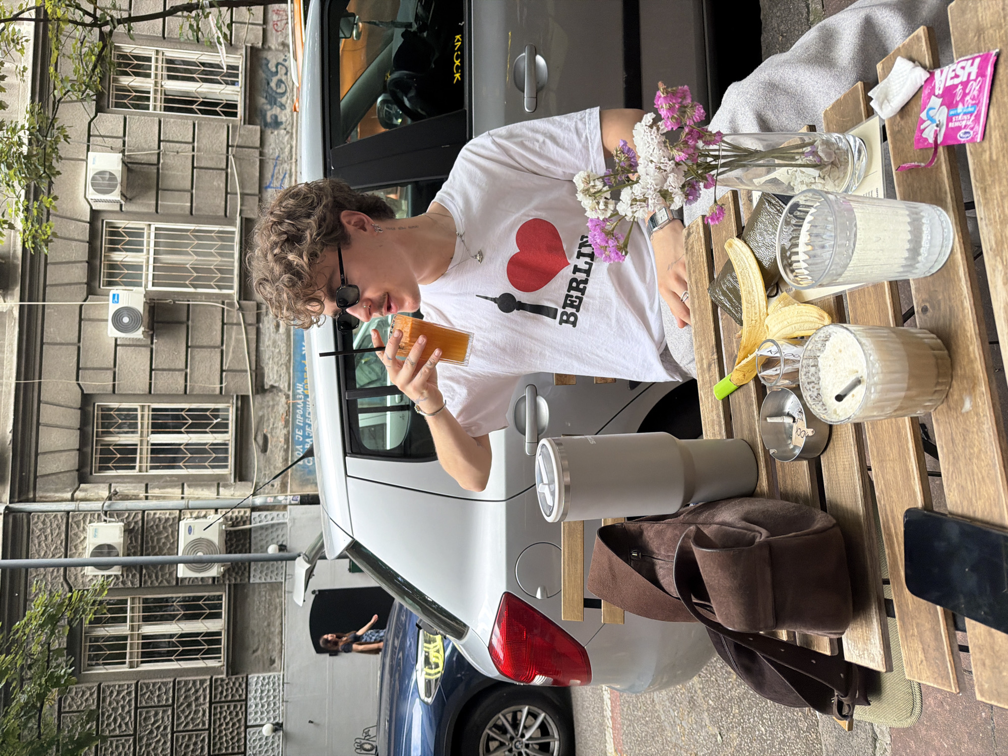 A young person with curly brown hair, wearing a white t-shirt with a red heart and "BERLIN" written on it, sunglasses, and jewelry, is drinking a beverage from a plastic cup with a straw. They are sitting at a wooden outdoor table next to a silver car.
On the table are: a vase with white and purple flowers, a glass of water, a peeled banana, a glass of a milky beverage, an ashtray with a cigarette butt, a grey thermos, a brown bag, and a black phone. There's also a small pink packet on the table.
The car is parked near a building with multiple windows and air conditioning units on its facade. Based on architectural style and street signs in Serbian, this photo was likely taken in Belgrade, Serbia. The orientation of the photo has been rotated.