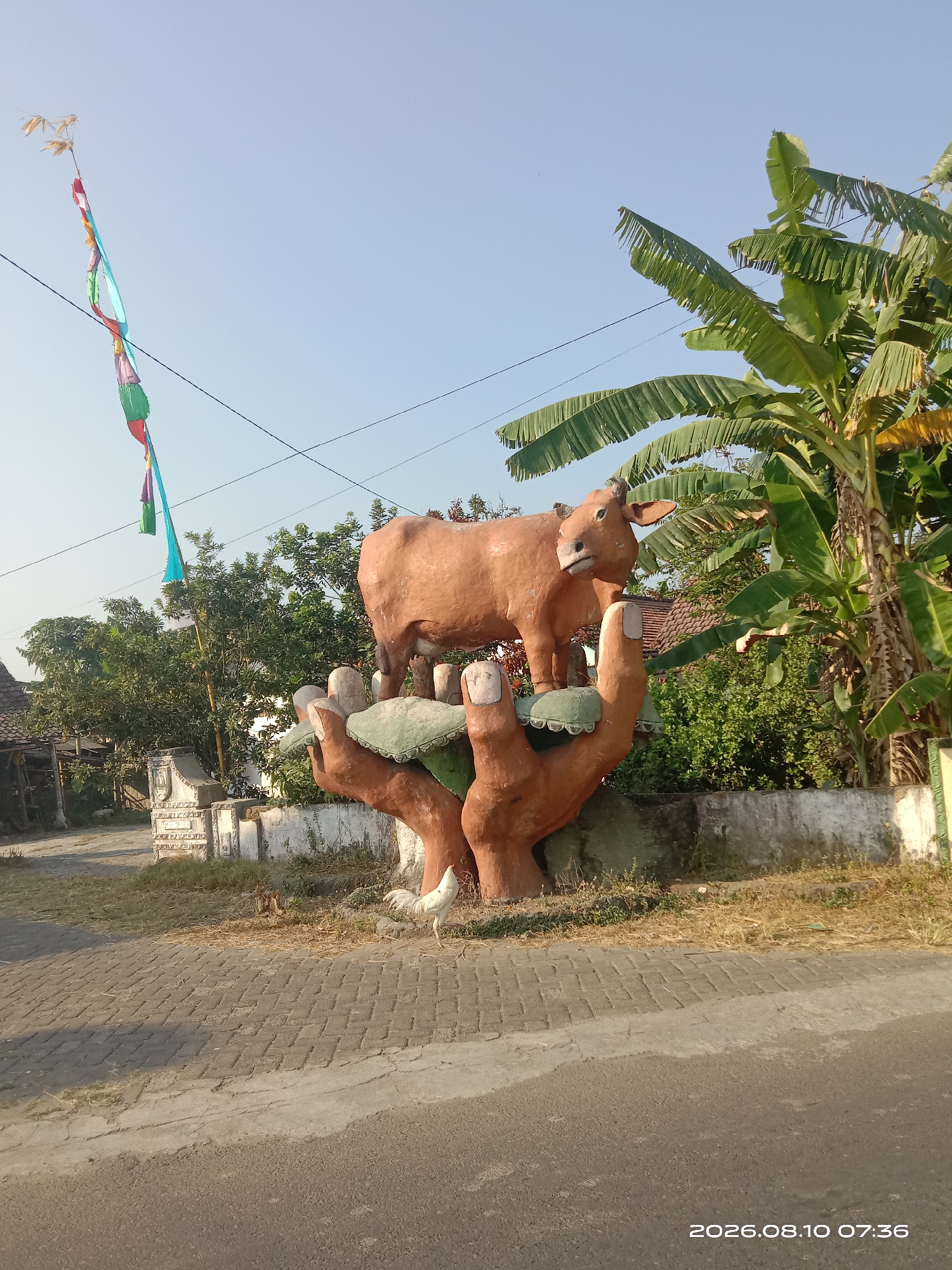 In Sidowarek, Indonesia, a statue of a brown cow stands on a large sculpture of two cupped hands, which are resting on the ground. The statue is made of concrete. The hands are textured to look like wood. The cow appears to be in a natural, standing position. The sculpture is located outdoors, with a clear blue sky in the background. Behind the statue, a cluster of trees and lush green foliage are visible, including banana trees with large, broad leaves. A white rooster is seen in front of the statue, pecking at the ground. The ground in front of the statue is paved with interlocking bricks and there is a road made of asphalt. A white decorative pillar is situated to the left of the statue. There are some buildings with tiled roofs in the background, suggesting a rural or semi-urban setting. Colorful streamers are attached to a pole to the left of the statue. The overall scene suggests a decorative roadside monument in a village or rural area.