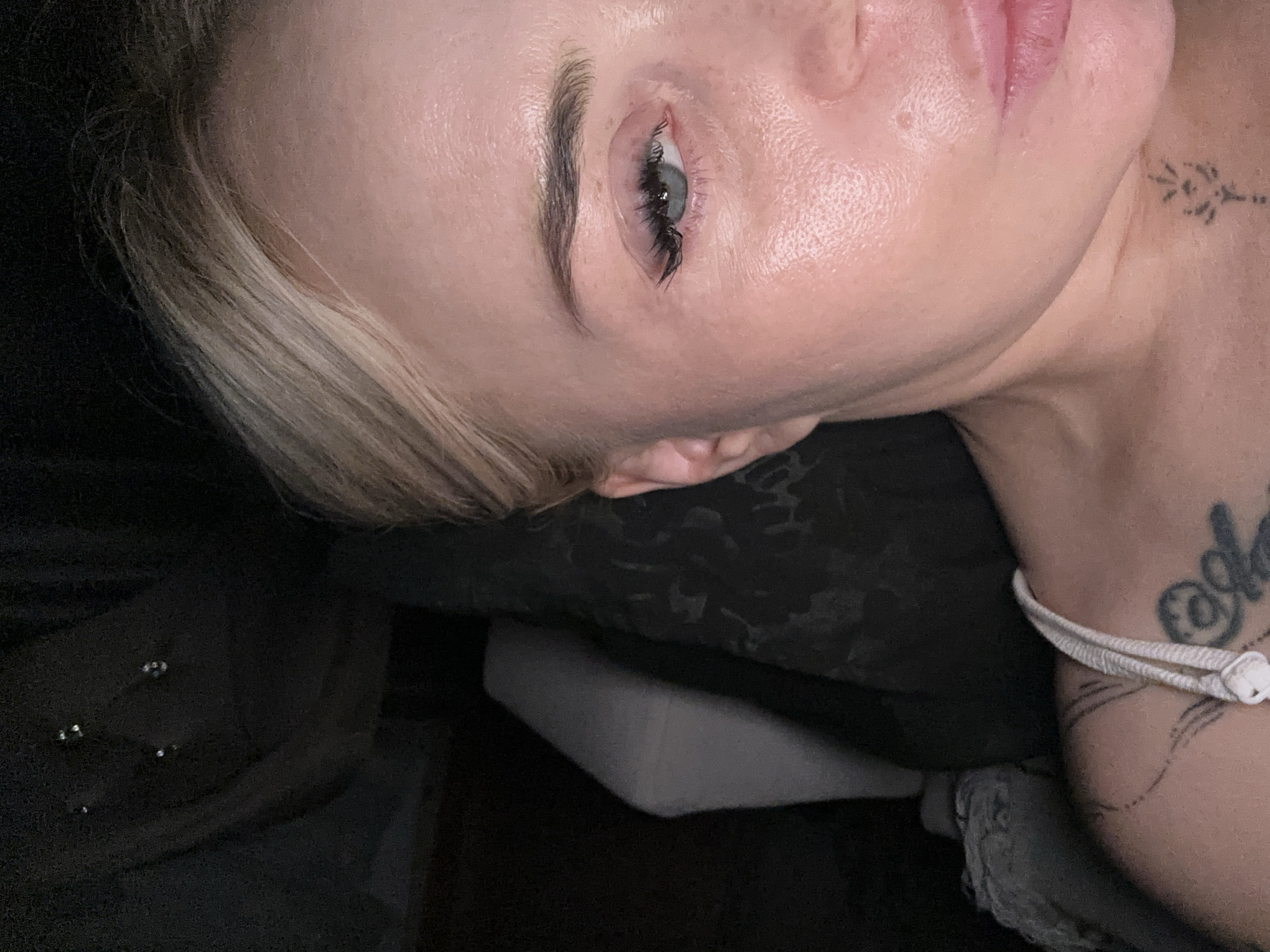 A close-up, vertically oriented photograph captures the right side of an individual's face and upper torso, set in a dimly lit environment in Unknown, Unknown. The subject is a fair-skinned individual with blonde hair, visible on the right, swept away from the forehead. The left eye (viewer's right) features a blue-grey iris, dark defined eyebrows, and prominent eyelashes with a winged appearance. The skin shows visible pores and a sheen, indicating moisture or reflective surface. The lower right portion of the frame shows the subject's full, light pink lips.
The subject wears a light-colored strap of a garment, likely a top or bra, on the left shoulder. Multiple tattoos are visible: a black line-art design on the upper left shoulder/chest, a smaller black linear symbol on the lower right neck/collarbone area, and a dark, partially visible tattoo featuring an "@" symbol and text on the chest beneath the shoulder strap.
The background on the upper left is dark and indistinct, showing several small, bright reflective specks. Below this, a grey, elongated object with a central crease, possibly a pillow or cushion, rests against a dark surface. The background directly behind the subject appears to be a dark, textured fabric, possibly upholstery. The illumination is consistent with artificial light, creating highlights on the skin.