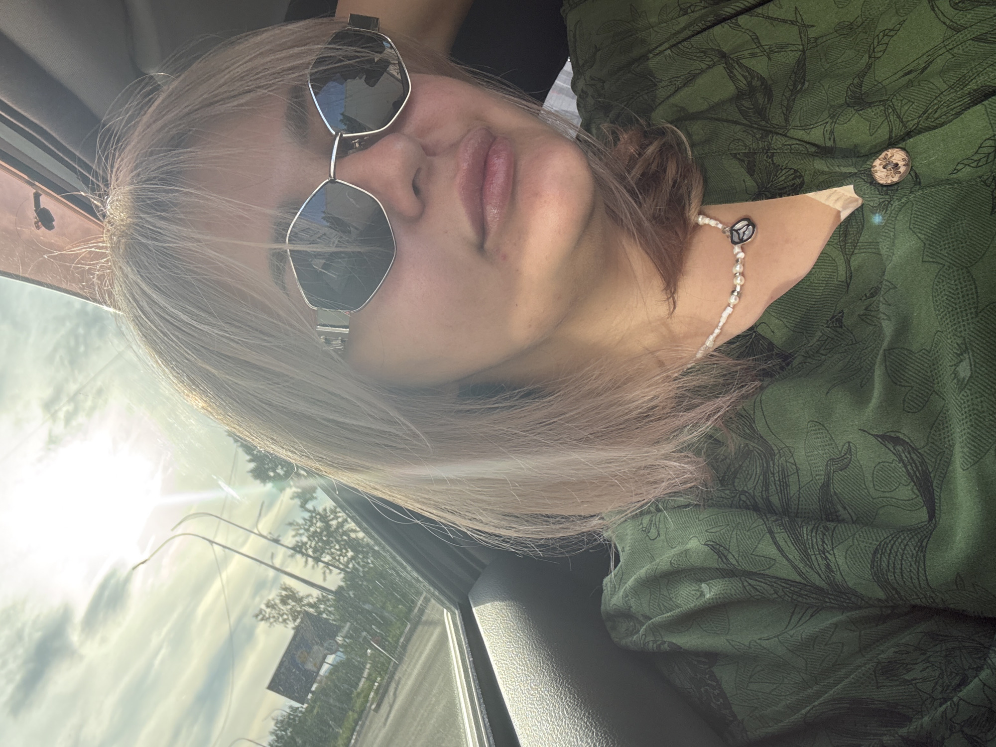 A woman with blonde hair and sunglasses is seated in the back of a car, looking at the camera. She is wearing a green, patterned shirt and a pearl necklace. The sun is shining brightly through the window, creating a lens flare. Outside the car, trees and a road are visible. The location is the city of Birobidzhan, Russia.