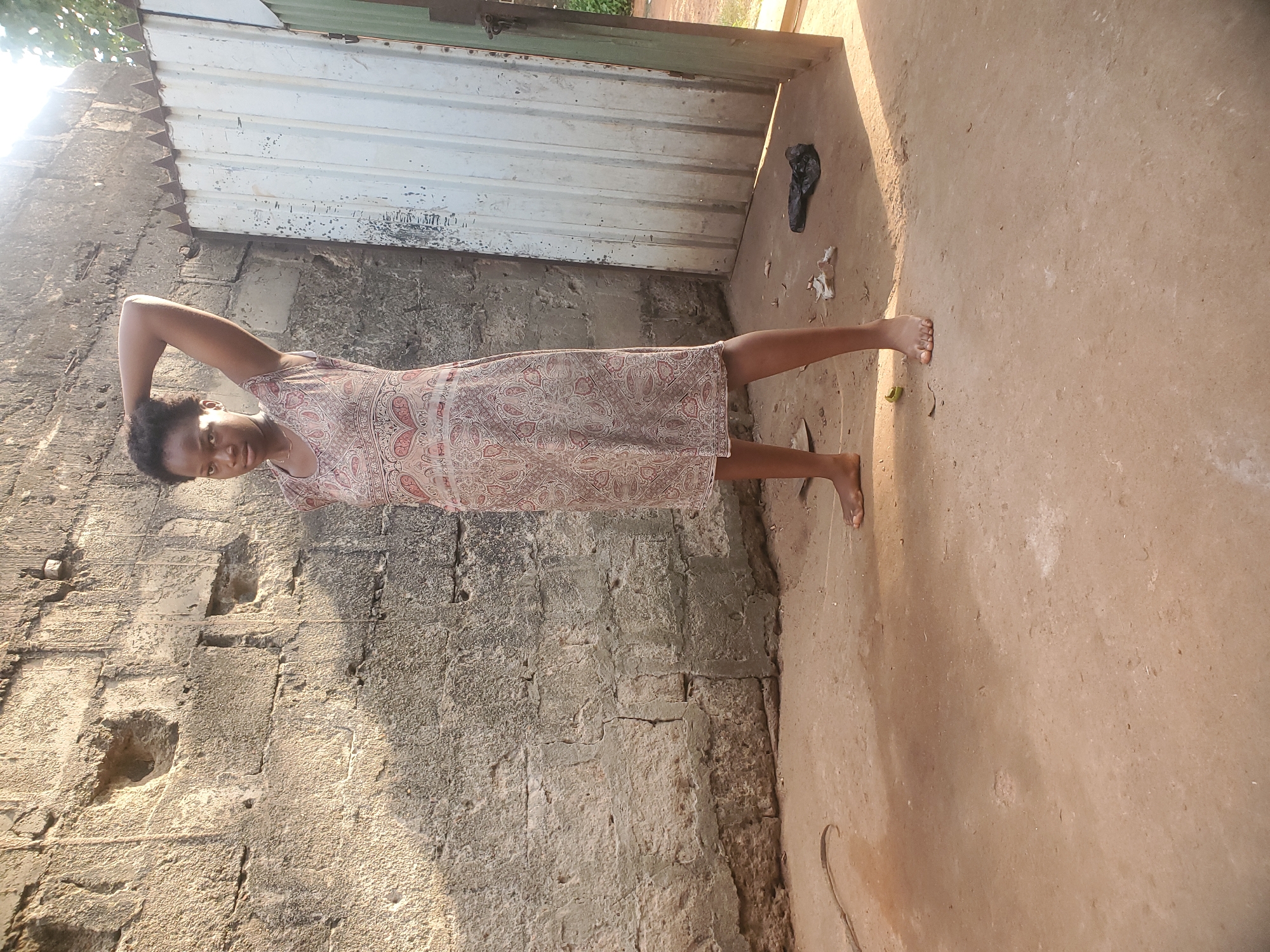 A young Black woman in a printed dress stands barefoot in Ukwu-Abwa, Nigeria. She faces the viewer, her left hand raised to her head. The background features a concrete block wall on one side and a concrete floor leading to a metal door on the other. A black object, possibly a bag or shoe, lies near the door. The lighting suggests an outdoor setting in daylight.