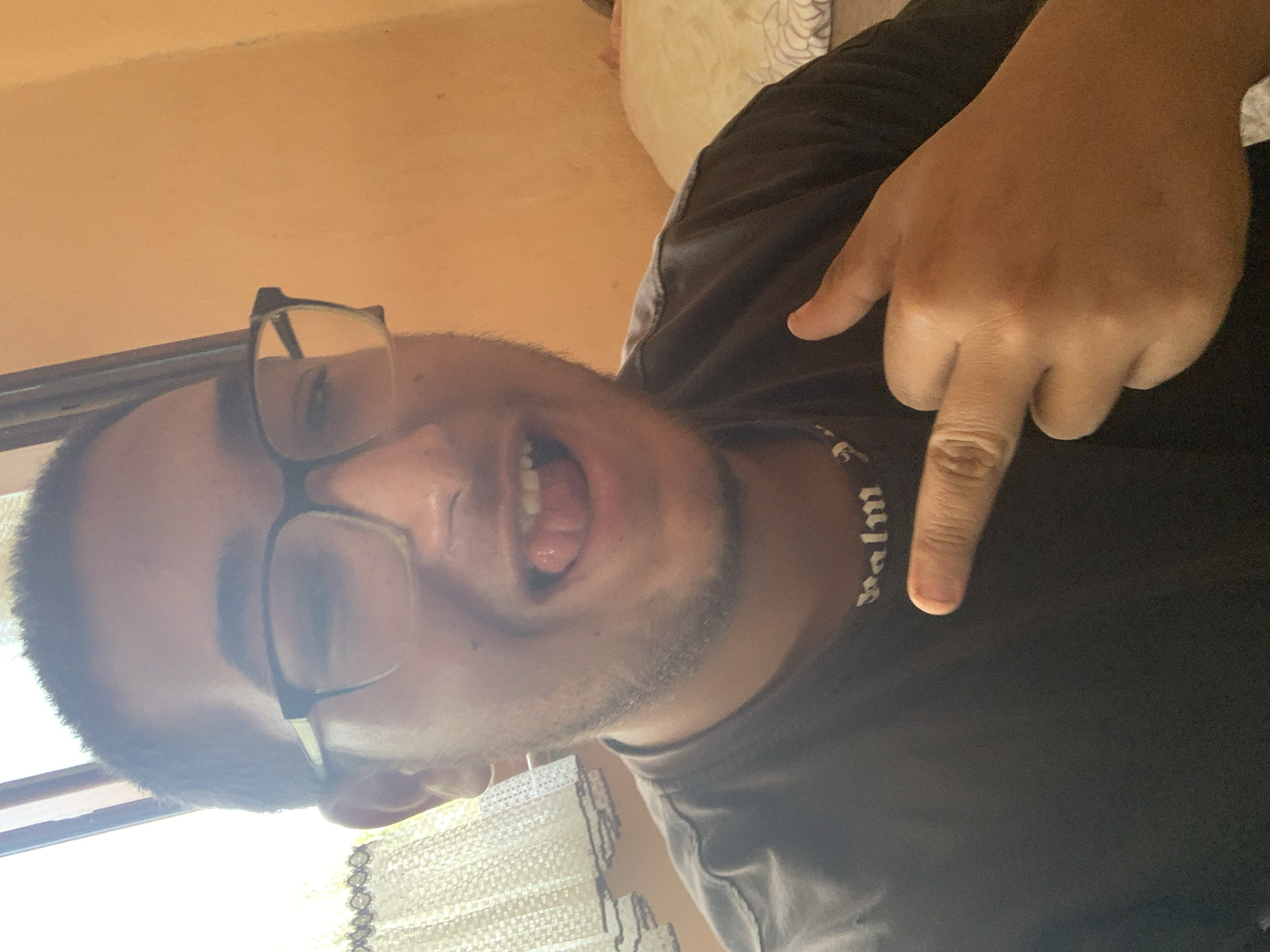 A person is shown in a medium shot, looking at the camera with their tongue sticking out. They are wearing glasses and a dark t-shirt, and a necklace with the word "PALM" is visible. The background consists of an orange wall and a curtain. The person is pointing their index finger towards the camera, and their right hand is partially visible. The overall lighting suggests it is daytime. The image is taken indoors.