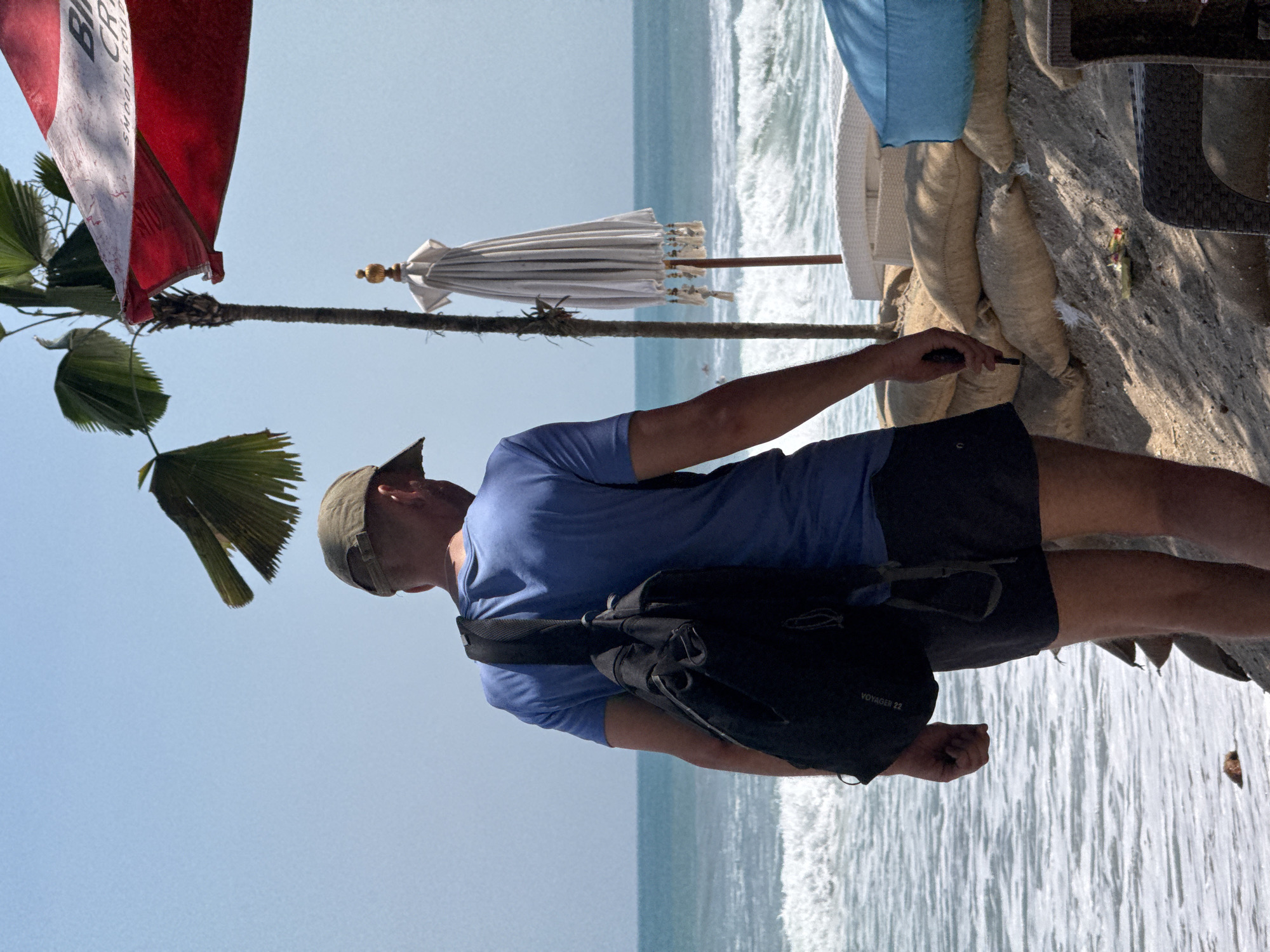 A man wearing a green baseball cap, a blue t-shirt, and black shorts stands facing away from the camera on a sandy embankment. He is wearing a black backpack. In the background, the ocean with small waves is visible under a clear blue sky. To the right of the man, a white umbrella with decorative tassels is propped up on a pole. A red and white striped umbrella is partially visible in the upper right corner. The embankment is reinforced with sandbags, and several white lounge chairs are arranged behind it. The location is Kuta, Indonesia, a popular beach resort town.