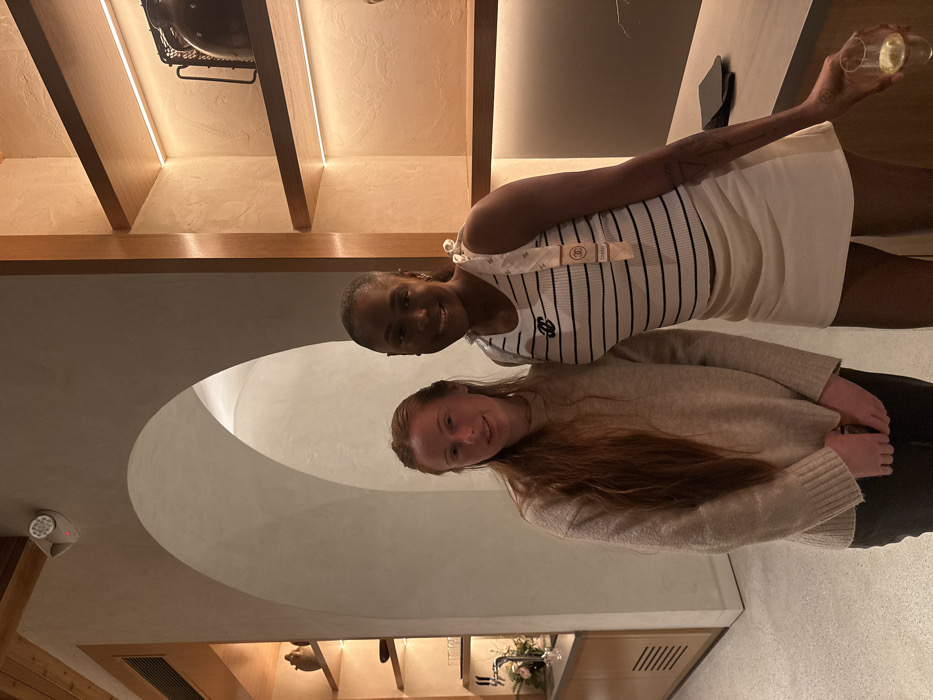 The image displays two adult females posing for a photograph within a modern commercial establishment, likely a restaurant or bar, located in the city of Los Angeles, United States of America.
The woman on the left is Caucasian, with long, straight, reddish-brown hair extending past her shoulders. She wears a light beige, long-sleeved knitted sweater and dark pants. Her hands are clasped at waist level. She looks directly at the camera with a slight smile.
The woman on the right is African American, with dark skin and dark hair pulled back from her face. She wears a white ribbed sleeveless top with thin black horizontal stripes and a black "Chanel" logo on the chest. A light-colored strap or scarf bearing "CHANEL" branding is draped over her right shoulder. She is dressed in a short, light-colored mini-skirt and dark hosiery. In her left hand, she holds a clear, stemless glass containing a pale yellow liquid. A geometric tattoo is visible on her left forearm. She is smiling broadly and looking directly at the camera. Both individuals are standing side-by-side, suggesting a posed interaction.
The background features smooth, light-colored walls and a prominent arched entryway in the center. To the left, a section of wooden shelving displays various decorative ceramic items and glassware above a counter with a small floral arrangement. To the right, a vertical section of similar wooden shelves, illuminated by strip lighting, holds decorative items including a dark urn with a handle. A dark, smooth countertop is visible beneath these shelves, with a black object resting on it. A white, cylindrical device, possibly a security camera or motion sensor, with a red light indicator, is mounted on the wall above the arched entryway to the left. The environment is characterized by warm, indirect artificial lighting.