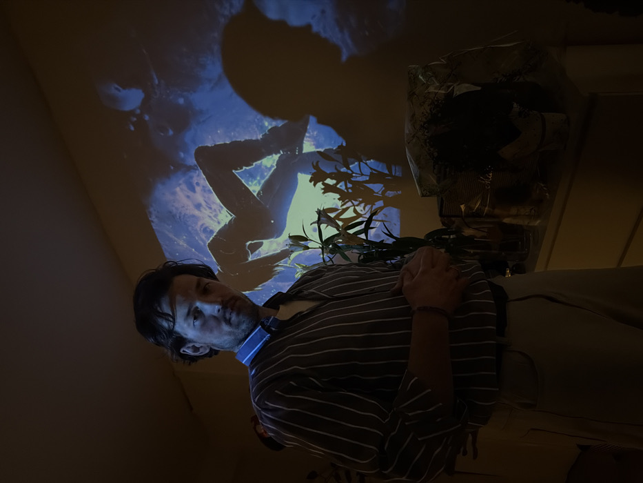 An adult male stands in a dimly lit interior room in Moscow, Russia. He is positioned centrally, facing forward, with his hands clasped at waist height. He wears a striped long-sleeved shirt with a visible blue collar and light-colored pants. A dark, beaded bracelet is on his right wrist. On the wall behind him, a blue-toned projection displays the silhouette of a human figure in a dynamic, possibly floating pose. To the man's right, a green plant with white lily-like flowers is situated in a clear vase on a surface. Next to the plant, a packaged item, partially wrapped in clear plastic, rests on the same surface. A distinct human shadow, visible in profile, is cast on the wall to the right of the projected image.