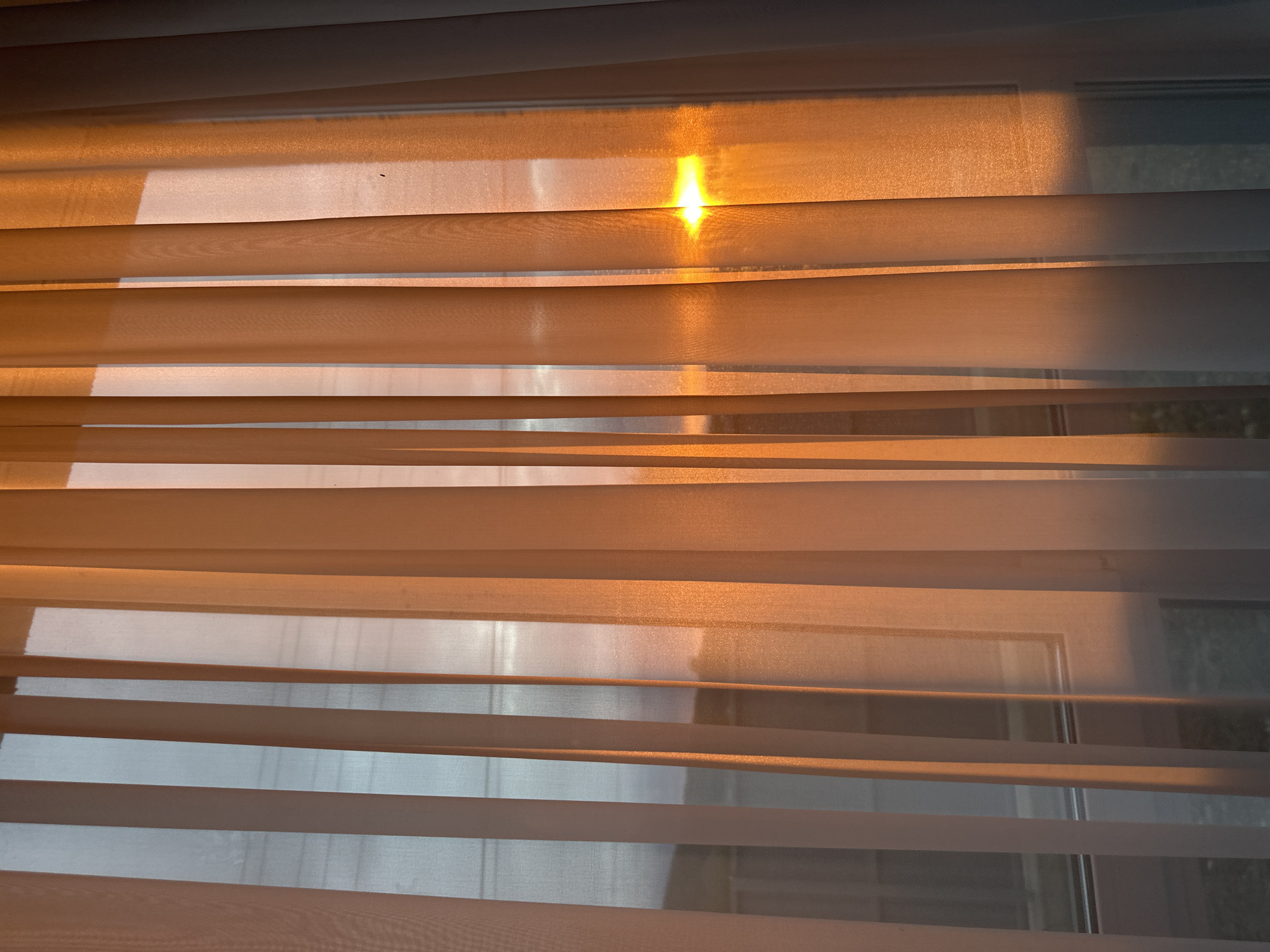 The media file displays an indoor scene featuring a window covered by horizontal blinds. These blinds appear to be a "zebra" or "day-night" style, characterized by alternating opaque and sheer fabric strips arranged horizontally across the window.
Warm, golden sunlight streams through the window, strongly illuminating parts of the blinds. A particularly bright, intense glow of light is visible towards the top center of the image, where the sun's rays directly hit the fabric, creating a radiant, star-like effect. The sunlight casts an orange and amber hue over much of the sheer fabric, while the opaque strips show varying shades of cream, beige, and orange, depending on how the light strikes them. Shadows from the opaque strips are visible on the sheer sections below, adding depth and texture.
Through the translucent parts of the blinds, a blurry outline of a window frame is discernable, along with indistinct shapes from the outdoor environment, possibly indicating foliage or distant structures, particularly in the lower right quadrant. The strong, low-angled golden light suggests either an early morning sunrise or a late afternoon sunset, contributing to a warm and peaceful atmosphere. No individuals or visible text are present in the image.