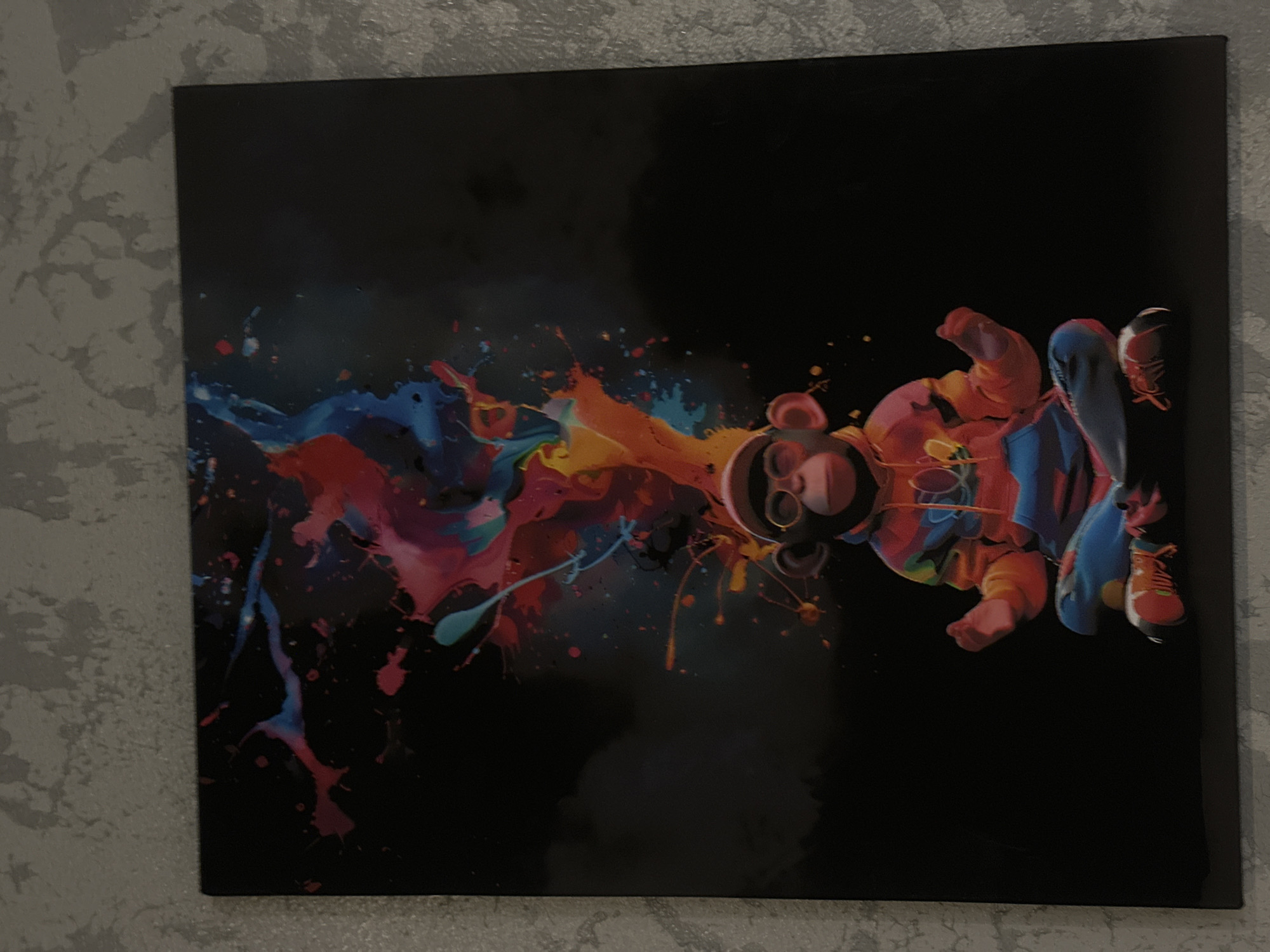 The image shows a canvas painting of a stylized teddy bear figure with paint splatters. The bear is predominantly orange and red, with blue accents on its clothing. It wears sneakers. The background is black, and splashes of colorful paint surround the bear, mainly in red, blue, and yellow hues. The canvas is hanging on a light grey wall. There is no indication of the location in the city of Chaikovsky, Russia.
