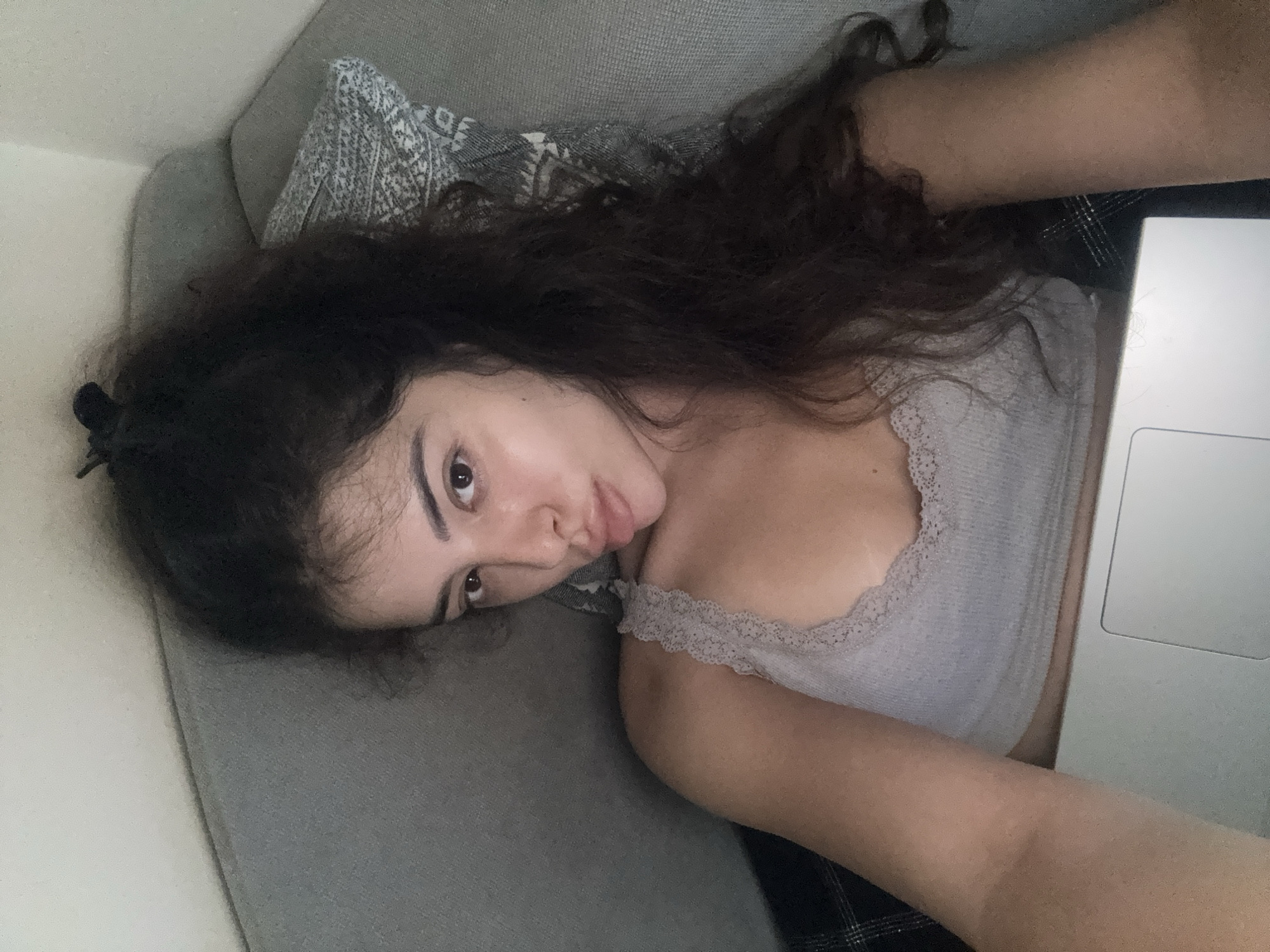 An overhead, close-up, selfie shot shows a young woman with long, dark, curly hair and fair skin. She is lying on a gray sofa with a patterned pillow and appears to be holding a silver laptop in her lap. The woman is wearing a light gray tank top with lace trim. Her lips are pursed, and her eyes are looking towards the camera. The background is a plain, light-colored wall. There is no indication of the location being Plovdiv, Bulgaria.