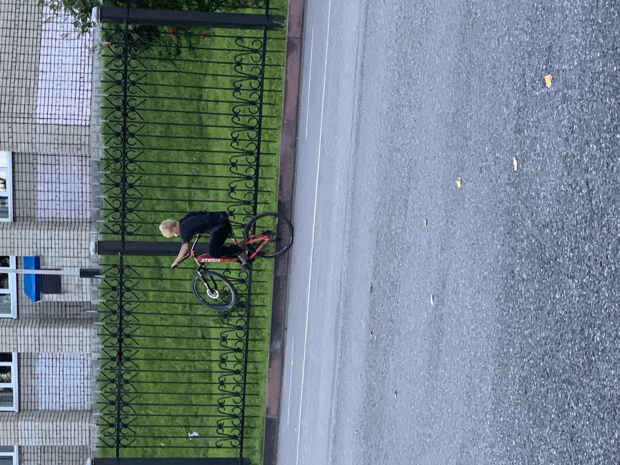 A young person with blond hair, wearing a black shirt and dark pants, is performing a wheelie on a red bicycle in Prokopyevsk, Russia. The cyclist is positioned on the asphalt road, with a decorative black metal fence and a patch of green grass behind them. Beyond the fence, a light-colored brick building with white-framed windows is visible. The scene appears to be outdoors on a cloudy day.