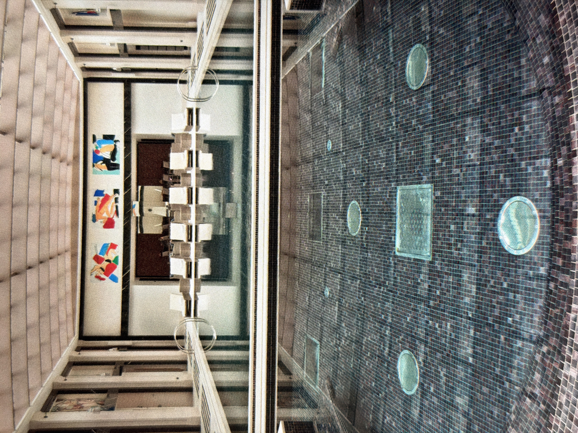 The image presents an interior shot taken from an elevated perspective, likely within a multi-story building located in Moscow, Russia. On the left side, there is a depiction of a hallway and staircase, featuring artwork on the walls and a contemporary lighting fixture. Adjacent to it, the right side displays a patterned tiled surface, possibly the bottom of a swimming pool, adorned with circular and square features. The architecture suggests a modern or post-modern design, with a blend of functional and decorative elements.