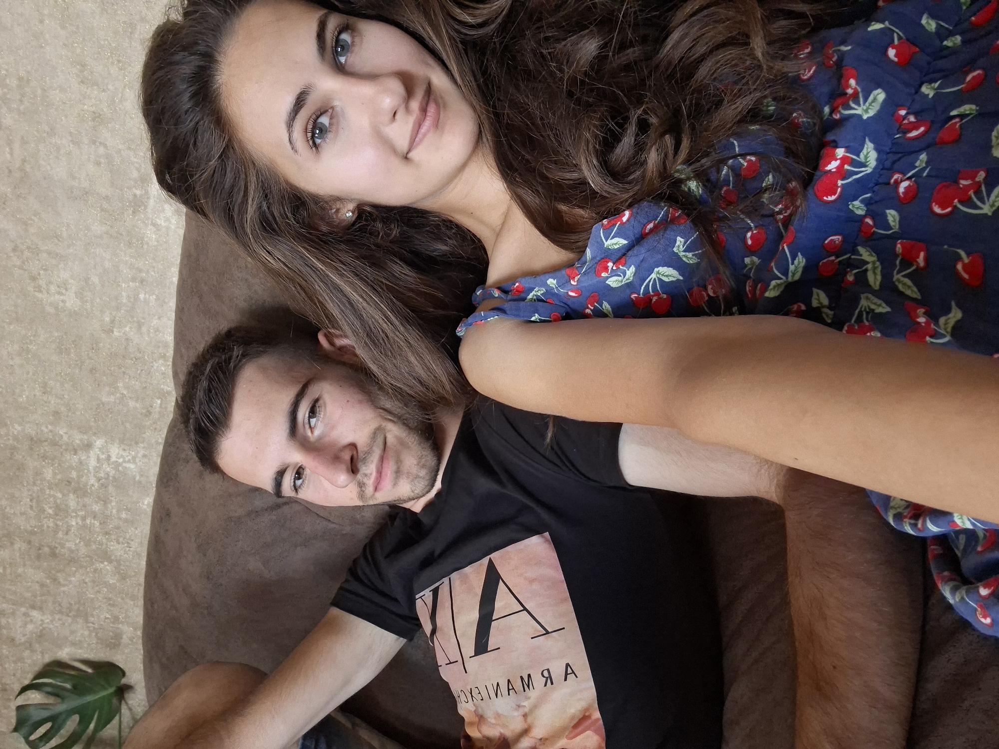 A young, dark-haired woman with blue eyes and fair skin is in the foreground, wearing a blue dress with a cherry pattern. Her hair is long and wavy, and she is smiling. To her left, a young man with short dark hair and a beard is lying down, wearing a black t-shirt with a graphic design. He is looking towards the camera. They are both lying on a brown couch. In the background, there is a wall with a textured pattern and a large green leaf from a plant. The scene appears to be indoors.