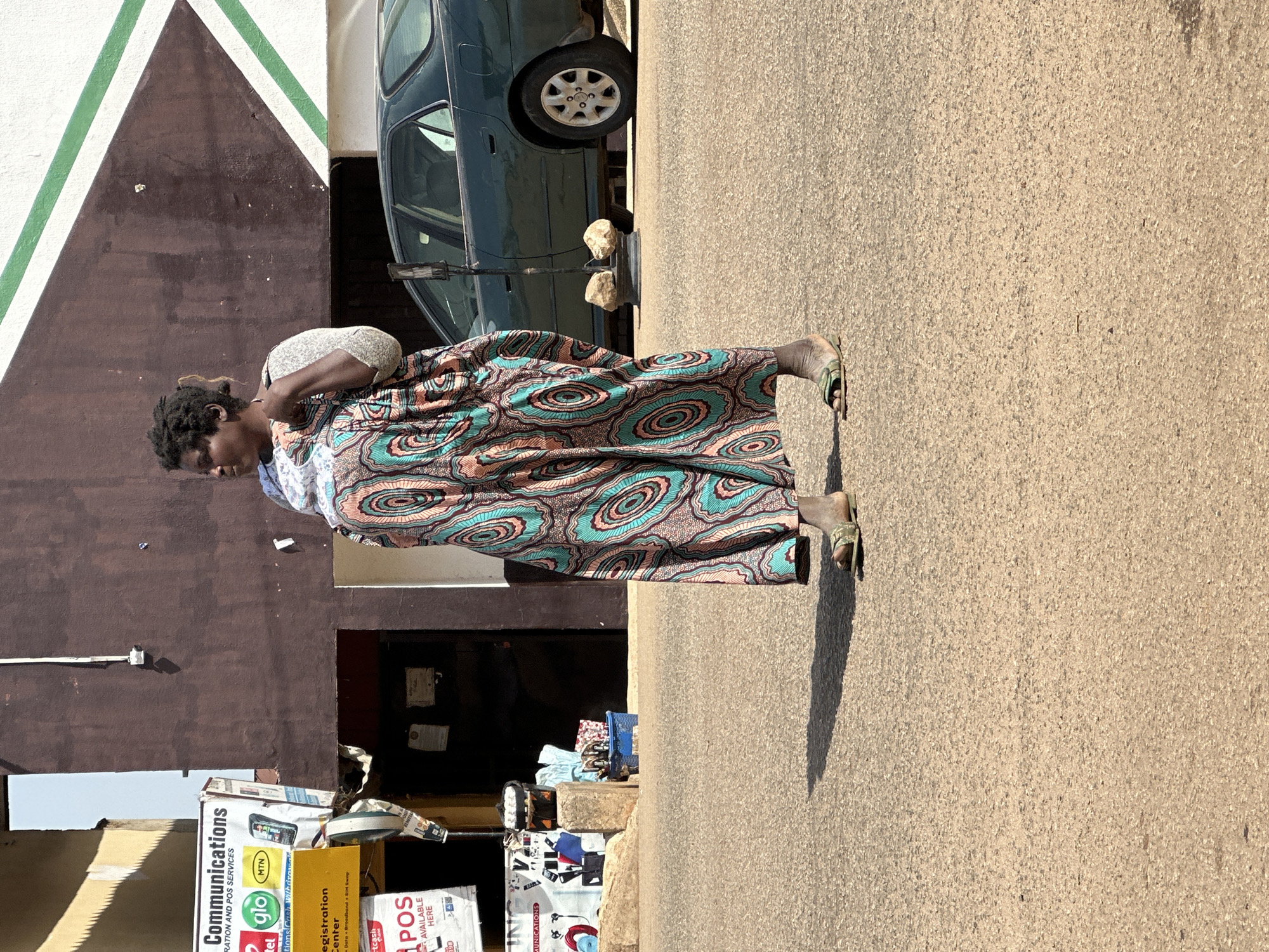 A full-body, eye-level shot shows a woman walking on a paved street in Bukuru, Nigeria. The woman is wearing a long, patterned dress with a light-colored fabric draped over her shoulder. She is walking from left to right, facing away from the camera. The background features a brown building with white and green geometric decorations on the upper portion. To the left of the woman, there is a yellow and white sign with text and logos, and a doorway. A dark green sedan is parked on the right side of the street. The ground is paved with asphalt and shows distinct shadows.