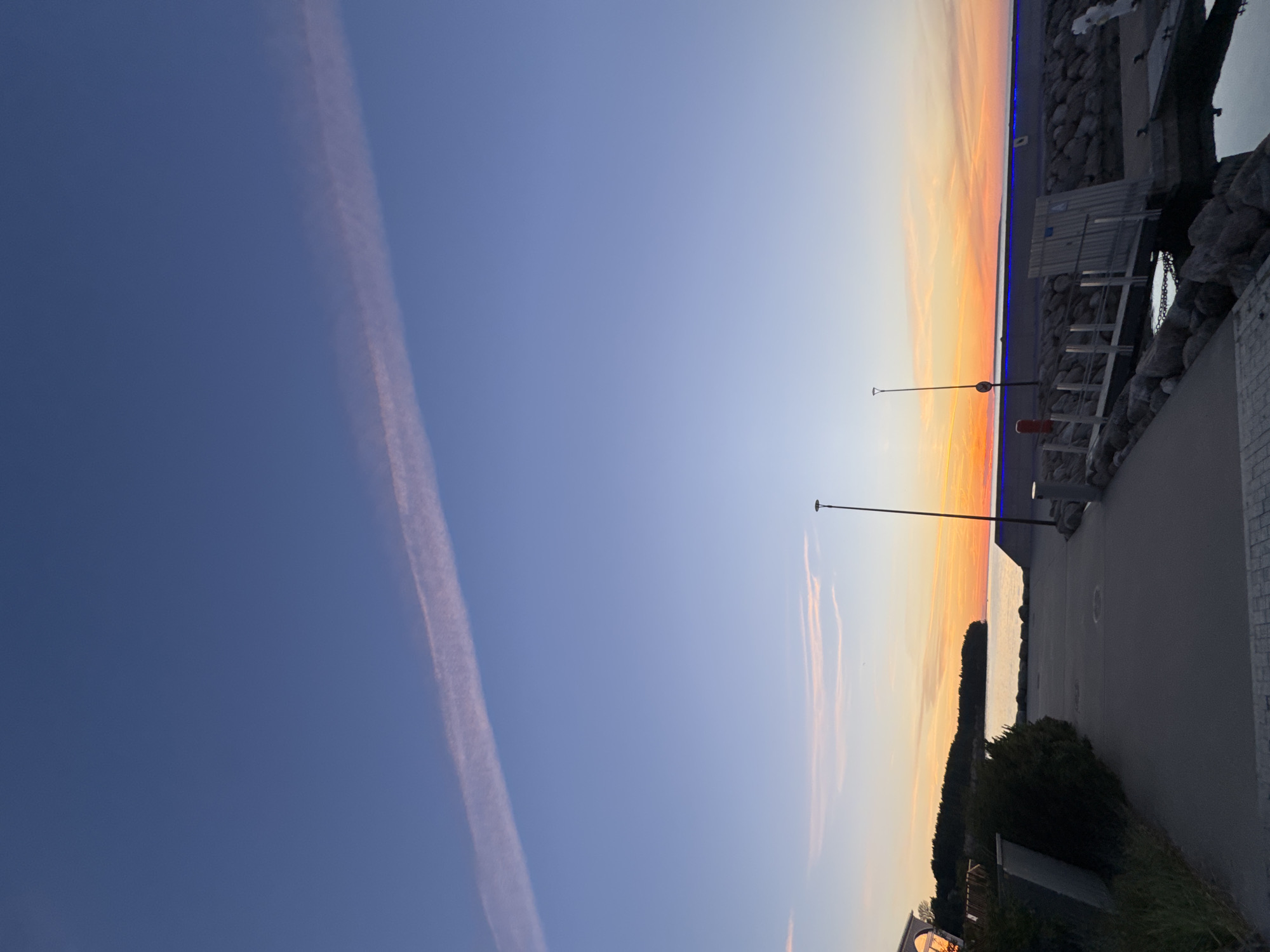 This is a landscape photograph of a harbor at dusk. The sky is a gradient of deep blue at the top, transitioning to pale blue and then a warm orange and pink near the horizon. A thin, wispy cloud stretches diagonally across the upper portion of the sky, catching the last light of the setting sun.
In the foreground, a paved walkway leads towards the water. To the left, there is lush green vegetation and a portion of a building with arched windows. To the right, there is a rocky seawall and a wooden pier extending into the calm water. Several lampposts are visible along the walkway and the seawall. The water reflects the colors of the sky, creating a serene and peaceful atmosphere. The scene appears to be a public space, possibly a marina or waterfront promenade. The location is not identifiable as Tallinn, Estonia from the visual information alone.