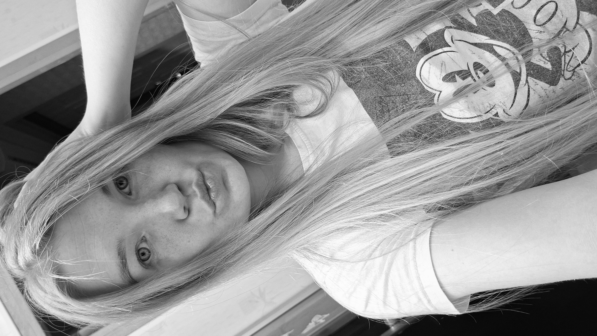 A black and white, close-up, selfie-style portrait shows a young woman with long, blonde hair partially covering her face and shoulder. She is wearing a t-shirt with a graphic of Mickey Mouse. Her right hand is raised, touching her hair. Her lips are pursed, and she is looking directly at the camera. The background appears to be indoors, with a wooden surface visible at the top. There is no indication of the city of Большой Букор, Russia, in this image.