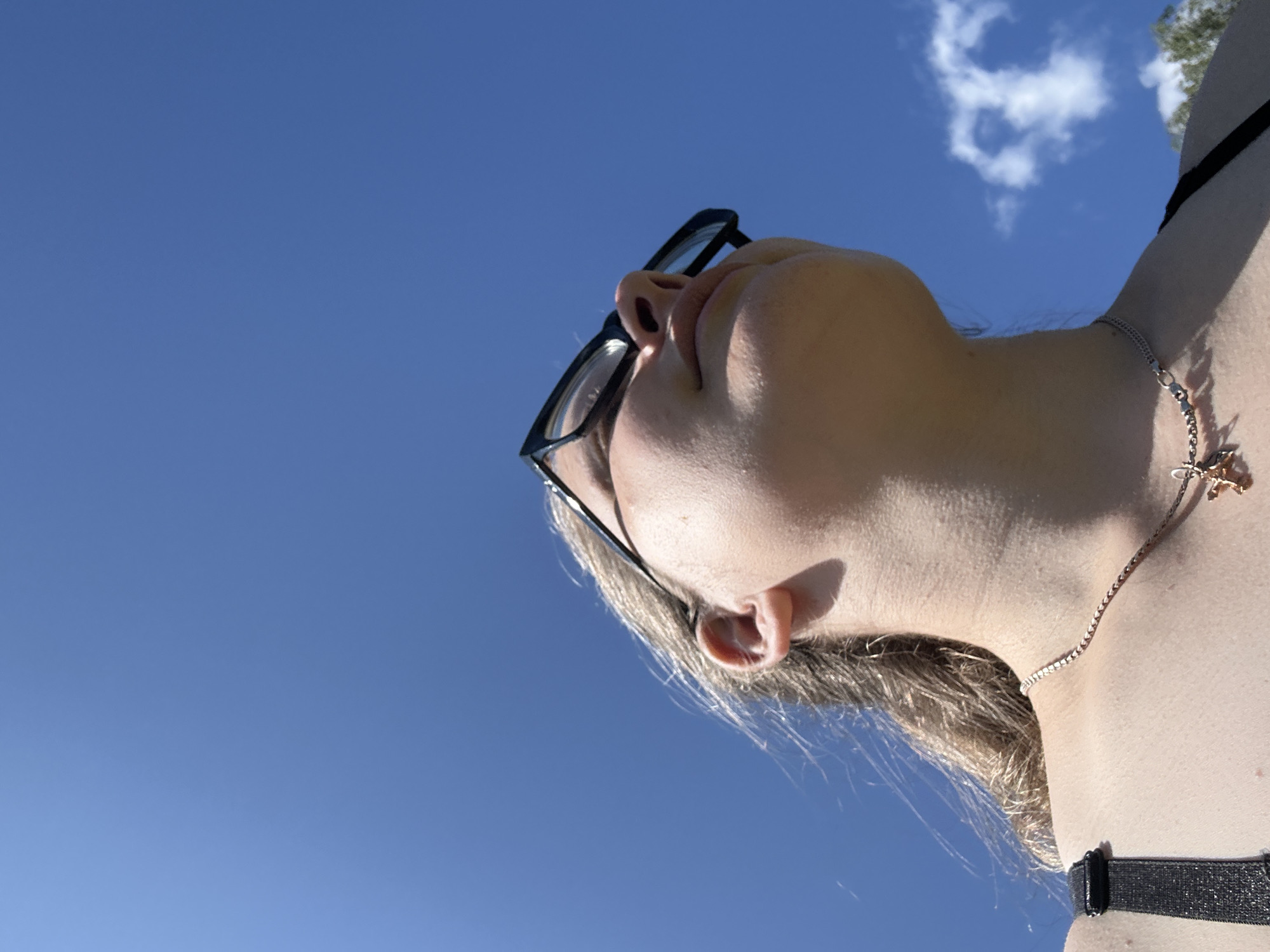 A low-angle, outdoor, full-body shot shows a person wearing black-rimmed glasses and a silver necklace with a small pendant. The person's blonde hair is windblown. The background is a clear blue sky with a few wispy clouds. A hint of trees is visible in the bottom right corner. The image is taken from the perspective of looking up at the person. The person's shoulders and chest are visible, suggesting they are wearing a tank top or a bikini top. The setting appears to be a sunny day.