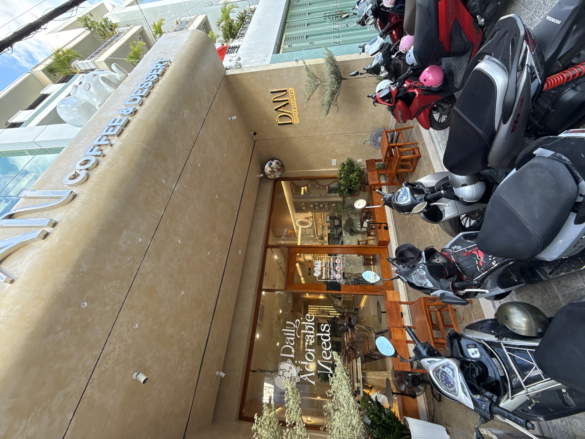 The image shows the exterior of a coffee shop named "DAN COFFEE & DESSERT" located in Nha Trang, Vietnam. The shop has a modern facade with a prominent beige awning featuring metallic lettering for the shop's name and "COFFEE & DESSERT". Above the awning, a white ceramic sculpture resembling a whimsical creature is visible.
The entrance to the coffee shop is through a glass door and large glass windows, allowing a view of the interior seating area with wooden tables and chairs. On the left side of the entrance, white lettering on the glass reads "Daily Adorable Needs" with a small illustrated mouse icon. To the right of the entrance, a neon sign displays "DAN COFFEE & DESSERT". A sign on the door indicates "Opening Hours Monday - Sunday 6:30 am - 22:00 pm" along with a series of circular icons, likely representing policies or amenities.
In front of the coffee shop, several motorbikes are parked. These include a black and silver scooter in the foreground on the left, a red scooter with a pink helmet hanging from the handlebars, and other scooters further back. The ground is paved with grey tiles. Green shutters are visible on the right side of the building.
The environment appears to be an urban street with other buildings in the background, including balconies with plants. The sky is blue with scattered clouds, suggesting daytime. The overall scene depicts a commercial establishment in a city, with a focus on the coffee shop's exterior and its immediate surroundings.