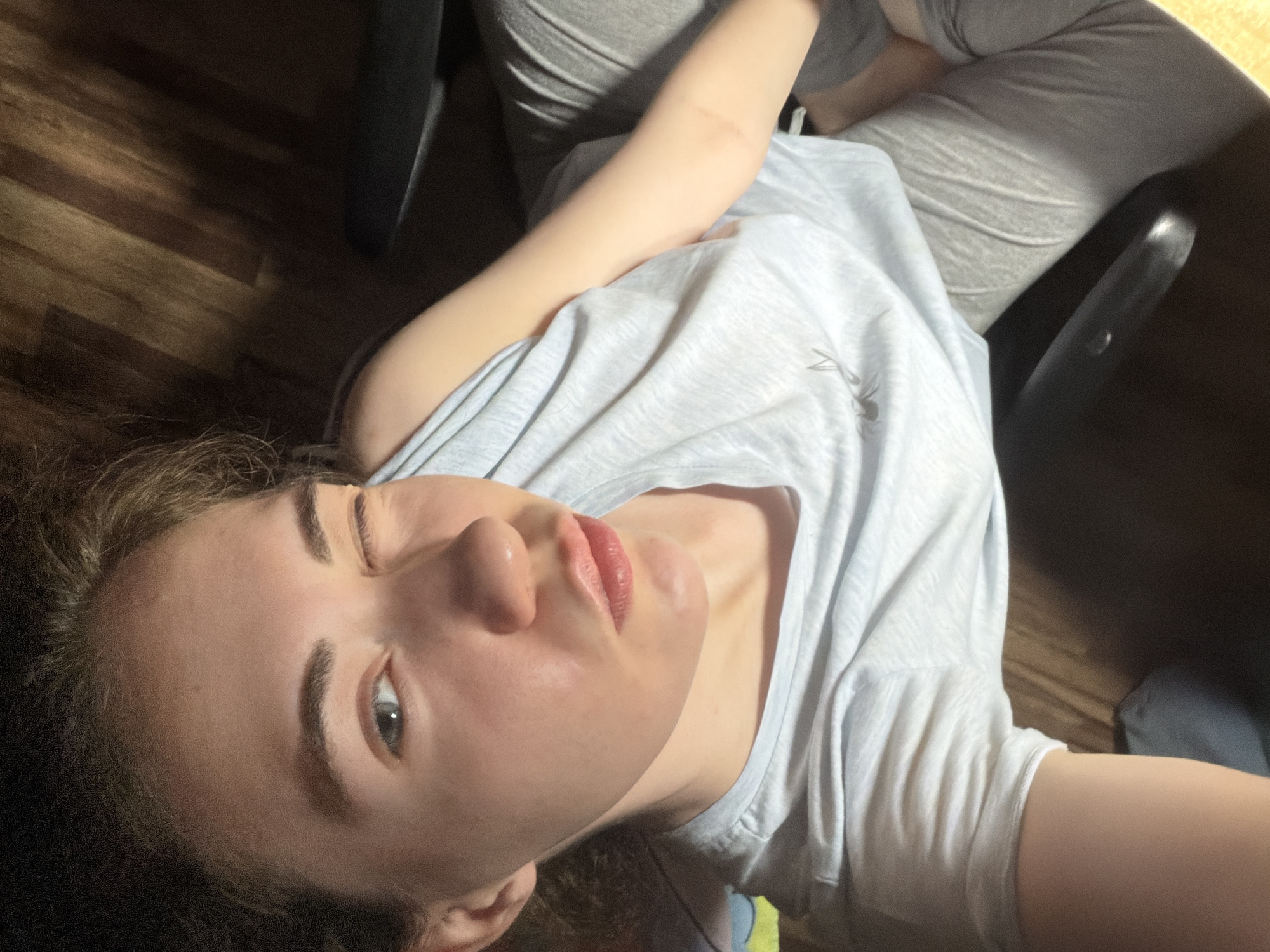 A high-angle, close-up, selfie-style photograph shows a young Caucasian woman from the chest up. She has brown hair pulled back and is wearing a light blue t-shirt and grey sweatpants. Her left eye is open and looking at the camera, while her right eye is closed in a wink. Her lips are slightly parted. The background consists of dark wood flooring and what appears to be furniture. No specific location in Zavodoukovsk, Russia, can be determined from the image.