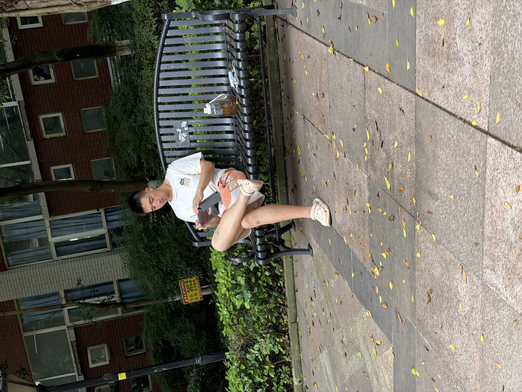 A young man sits on a black metal park bench in Chongqing High-tech Zone, China. He wears a white t-shirt and orange shorts and rests his legs crossed at the knees. He is holding a tablet. A clear water bottle and a blue mask are on the bench. The ground is tiled concrete with scattered yellow leaves. A lamp post, green bushes, and brick buildings with windows surround the bench.