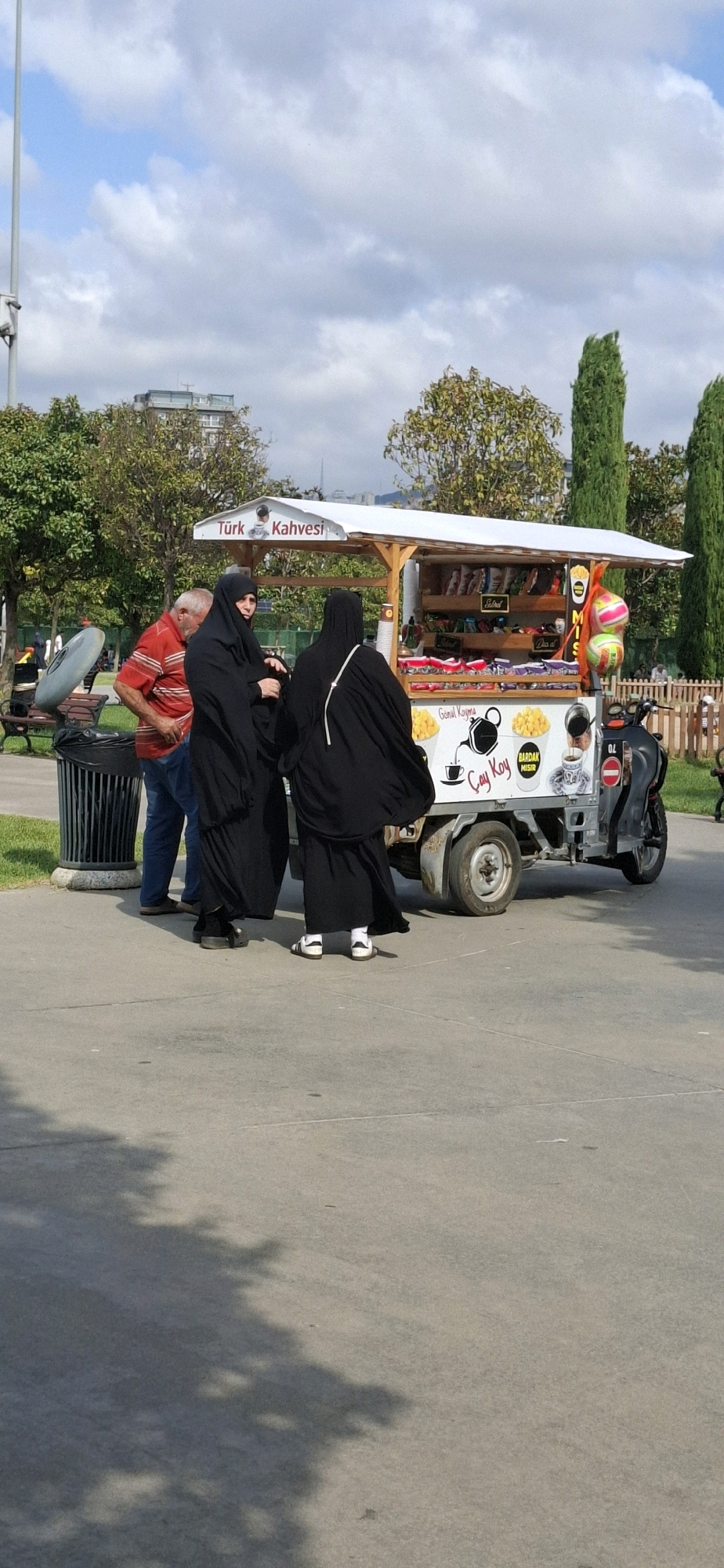 A mobile food cart is parked on a paved surface in an outdoor setting, possibly a park or public walkway. The cart has a white canopy and is decorated with colorful images and text. One side of the cart displays "Türk Kahvesi" (Turkish Coffee) above an image of a man's face. Below this, there are shelves stocked with bags of snacks and other packaged goods. The lower portion of the cart features graphics of a teapot, cups, and text that includes "Çay Koy" (Put Tea), "Gönül Koyma" (Don't Get Upset), and "Bardak Mısır" (Cup Corn). A small motorcycle is attached to the rear of the cart.
Three individuals are present near the cart. Two women are dressed in black abayas, covering their hair and bodies. One woman, facing the cart, has her hands clasped. The other woman, partially obscured, is facing away from the camera. A man in a red and white striped shirt and blue jeans stands to the left of the women, looking towards the cart.
In the background, there are trees with green and yellow foliage, indicating it may be autumn. A tall, slender pole with what appears to be a light fixture is visible to the far left. A dark cylindrical trash bin with a gray lid is situated to the left of the people. The ground is a light gray paved surface with shadows cast by trees. The sky is partly cloudy with blue patches.
The context provided suggests this scene is located in Maltepe, Turkey.