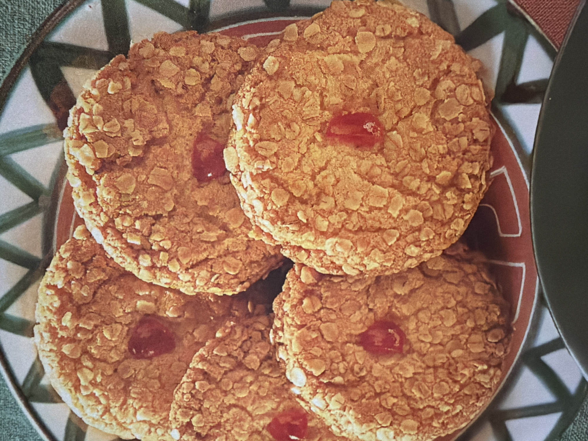 The image shows a close-up of several round, golden-brown cookies, possibly oatmeal cookies, arranged on a decorative plate. The cookies have a textured surface with visible oats and appear to be topped with small, bright red pieces, likely cherries or fruit preserves. Four cookies are prominently featured, with portions of others visible. The plate has a pattern of green and white stripes interspersed with red accents. The background is softly blurred, suggesting a focus on the food. The lighting is warm and diffused, creating a cozy and inviting atmosphere, possibly suggesting a domestic setting like a kitchen or dining area. There is no visible text in the image. The scene appears to depict a moment of enjoying or presenting baked goods.