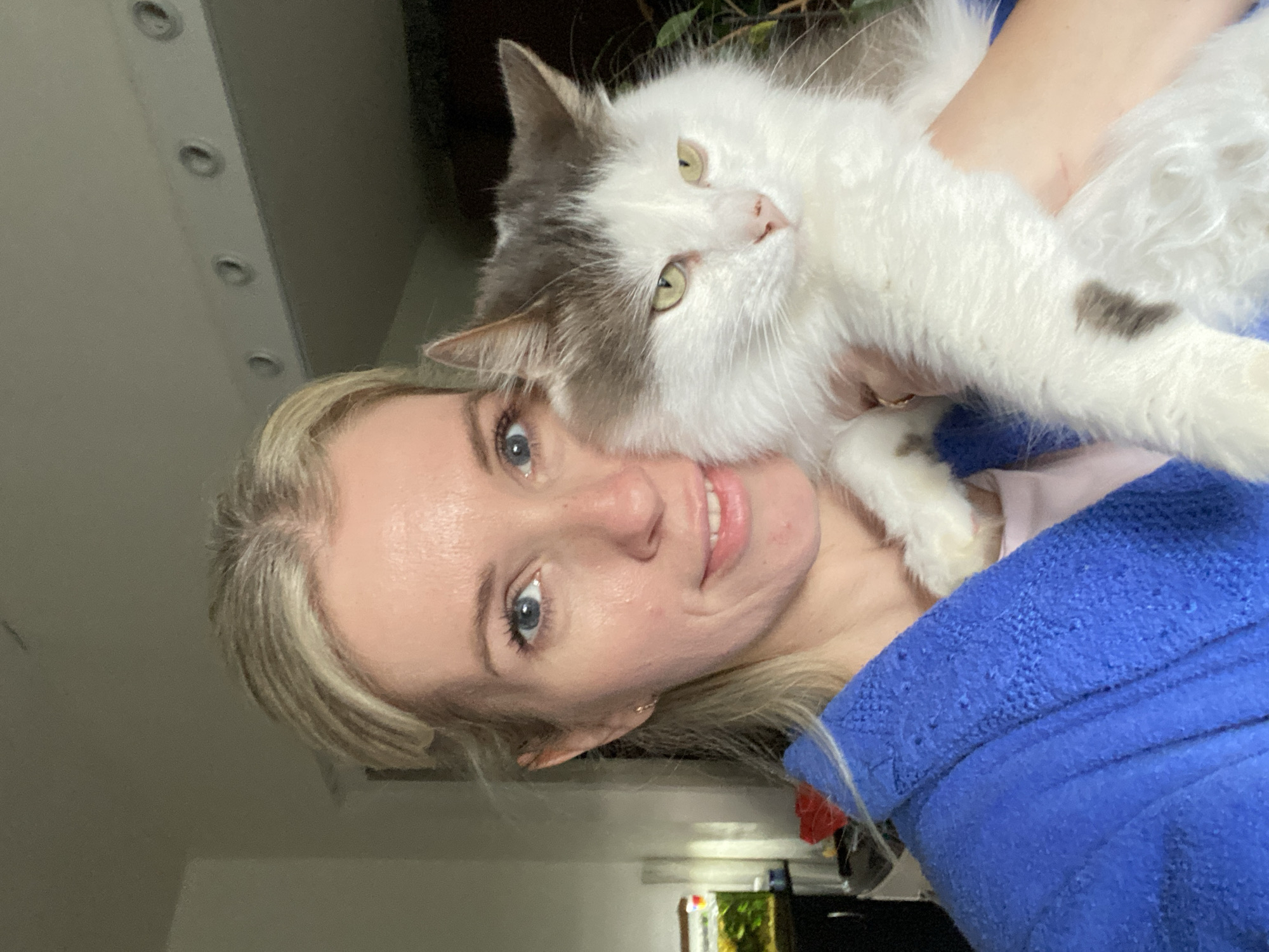 A woman with fair skin and light blonde hair is holding a fluffy white and grey cat. The woman is smiling and looking at the camera. She is wearing a blue robe. The cat is looking off to the side with yellow-green eyes. The background is slightly blurred, but shows part of a room with a ceiling that has embedded lights. There is also the top of what appears to be a fish tank to the bottom right. The location is Unknown, Unknown.