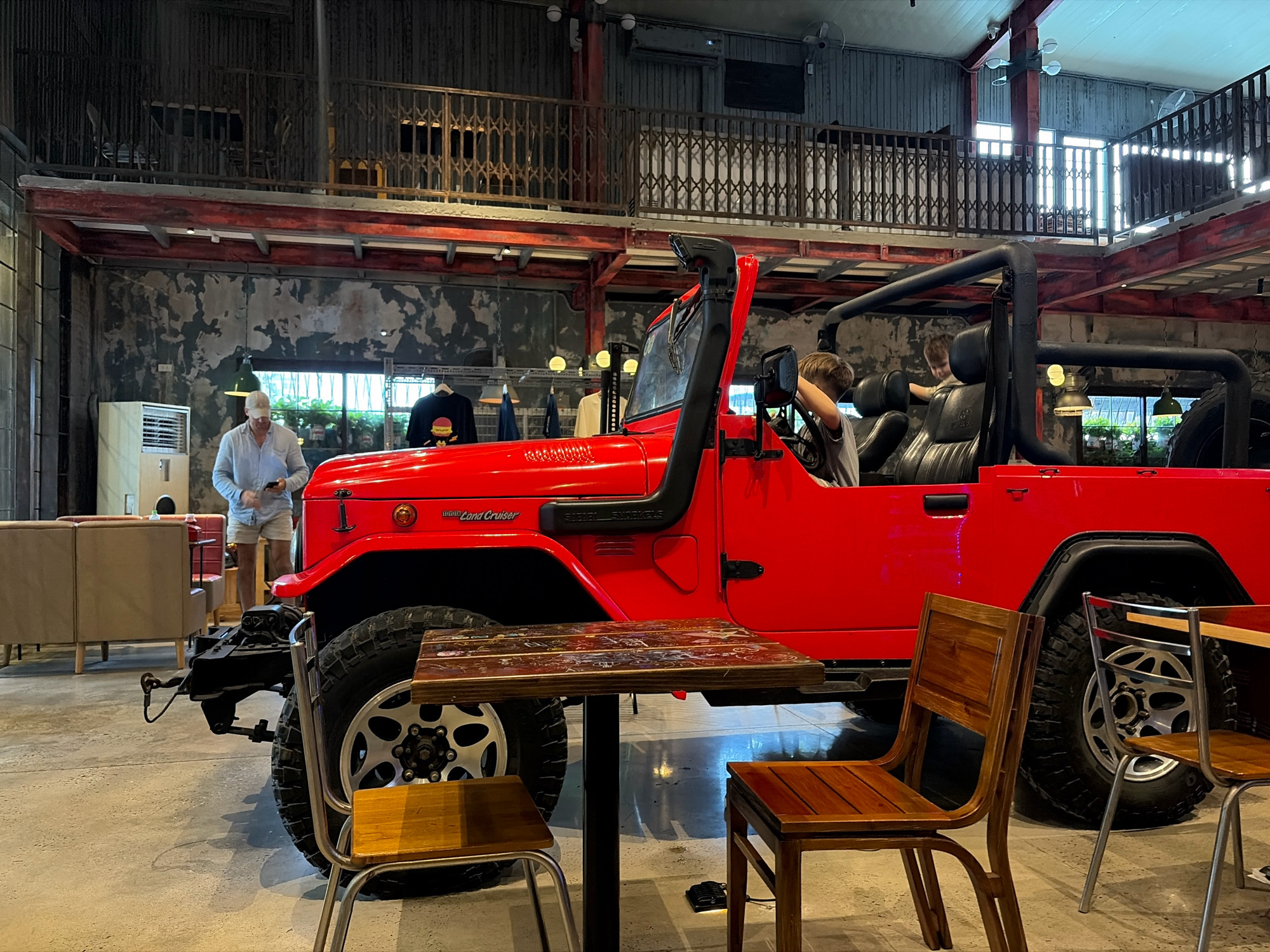 The image depicts the interior of a restaurant or cafe in Da Nang, Vietnam. The primary focus is a bright red Toyota Land Cruiser FJ40 parked indoors, serving as a unique seating area. Two young individuals, likely children, are visible inside the vehicle, with one appearing to steer. A man in a light blue shirt and shorts stands to the left of the vehicle, looking at his phone. Several tables and wooden chairs are arranged around the vehicle, suggesting a dining or lounge setting. The industrial-style interior features exposed concrete walls, metal beams, and a mezzanine level with railings. Natural light streams in from large windows in the background. The overall ambiance suggests a trendy and unconventional establishment.