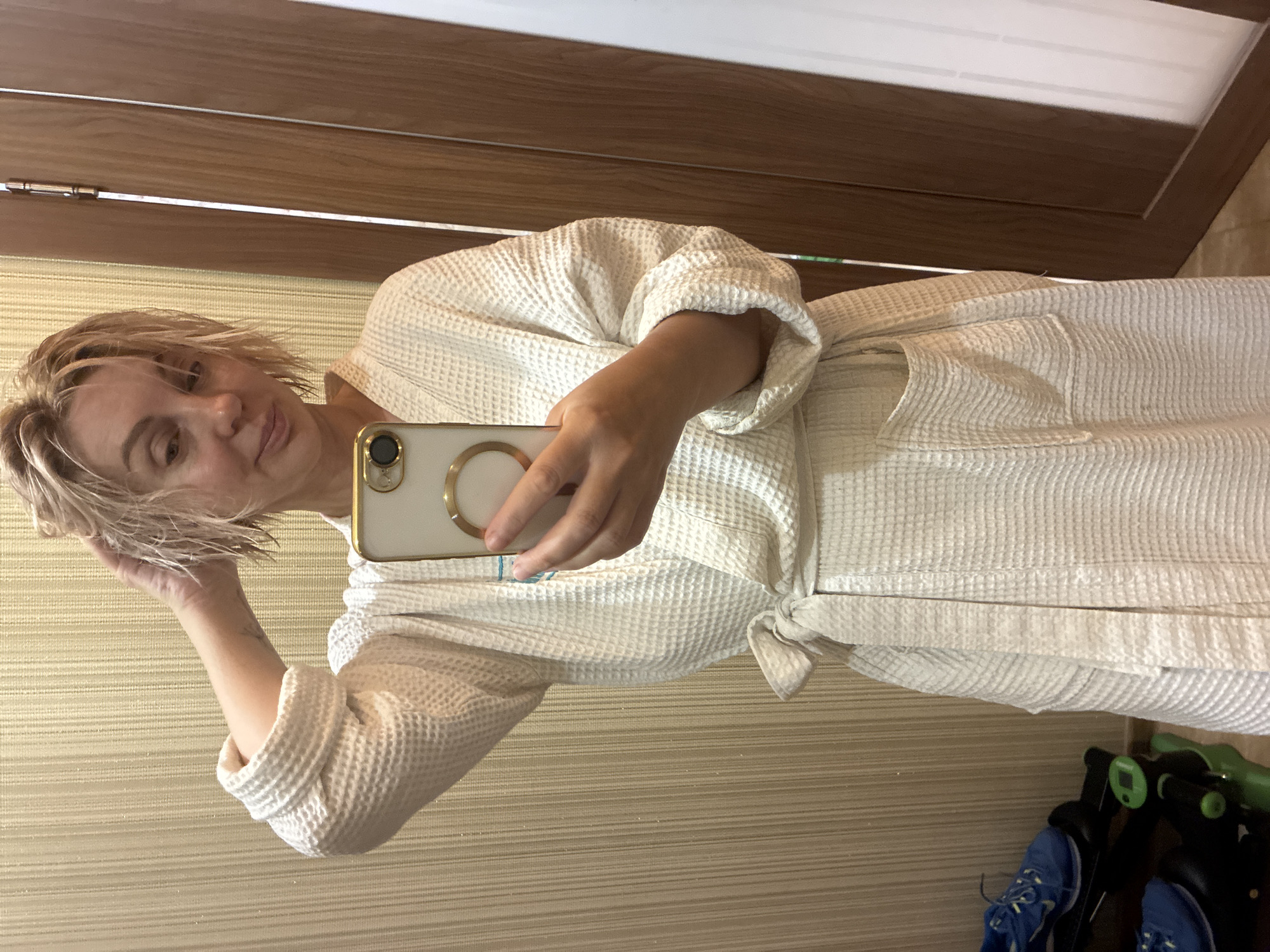 In a hotel room in Sochi, Russia, a woman with wet blonde hair, wearing a white waffle-knit bathrobe, takes a selfie in a mirror. She is holding a gold-trimmed smartphone in her right hand, and her left hand is in her hair. Her left arm is bent at the elbow, and her hand is touching her head. The bathrobe is tied at the waist with a white sash. In the background, a wall with a textured, striped wallpaper is visible, along with a wooden door frame and a glimpse of a doorway. To the left of the woman, a pair of blue athletic shoes are visible on a black and green rack. The lighting appears to be artificial indoor lighting.