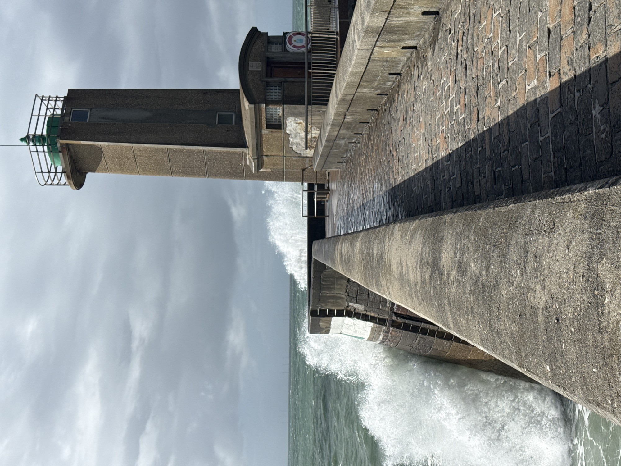 This vertical image captures a robust concrete lighthouse and an accompanying pier or breakwater at Fécamp, France, amidst rough sea conditions on an overcast day. The perspective is low, looking up at the towering structures from the water.
In the midground, a tall, rectangular concrete lighthouse dominates the scene, featuring a green cap and a protective metal cage structure at its apex. Small windows are visible on its shaft. Adjacent to the lighthouse, a smaller concrete building, integrated into the pier, has a rounded section, a visible wooden door, and what appear to be glass block windows. A red and white lifebuoy hangs on its exterior wall.
The substantial concrete pier extends from the foreground into the distance. Its visible side features a textured, brick-like wall, with a distinct, sharp shadow falling across part of its surface, hinting at a strong but possibly obscured light source. Powerful white waves crash vigorously against the base of the lighthouse and the pier, generating considerable spray and foam. Metal railings and ladder elements are also visible along the pier structure.
The sky above is heavily overcast with light grey, textured clouds, contributing to the dramatic and slightly muted lighting of the scene. The sea beyond the immediate wave action appears turbulent and dark green-grey. The overall impression is one of the enduring strength of the man-made structures against the raw power of the ocean. No people or discernible text are visible.