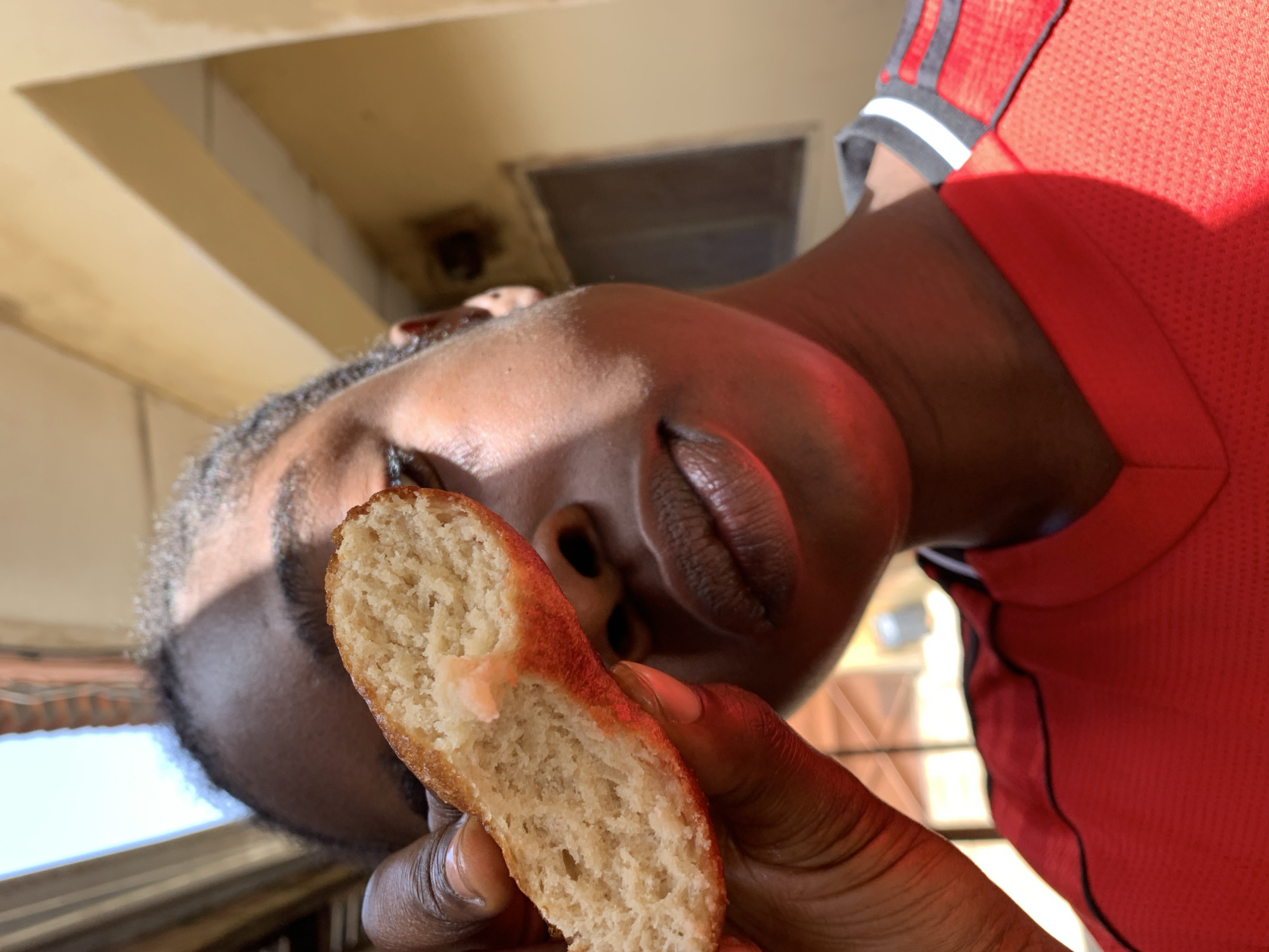 A close-up, eye-level, outdoor shot shows a young Black person holding a piece of fried dough in front of their face, obscuring the left eye and part of the forehead. The person is wearing a red shirt with black and white trim on the collar and sleeves. The background is out of focus, with a light yellow wall and a segment of a window visible. Sunlight casts shadows on the person's face. The setting appears to be a residential area in the city of Rafin Par, Nigeria.