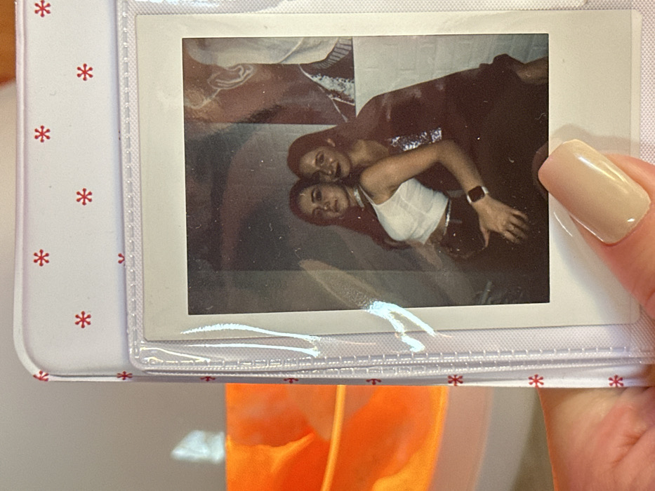 The scene depicts a human hand holding a white photo album page in 3-Y Uchastok, Russia. The album page features a repeating pattern of small red asterisk-like symbols and contains a single instant-print photograph secured within a clear plastic sleeve. The hand, with a short, nude-polished fingernail, grasps the bottom right corner of the album.
The instant photograph, a square-format print with a white border, displays two young adult females posing indoors. The female on the left has long dark hair, wears a white sleeveless top, a silver choker, and dark trousers. A dark smartwatch is visible on her left wrist, and her left hand rests on her hip. She looks directly at the camera with a neutral expression. The female on the right, also with long dark hair, is positioned slightly behind and to the right of the first subject. She wears a dark, possibly sequined top and has a slight smile or smirk. Her right arm is wrapped around the first subject's waist, with her hand resting on the left subject's left hip/buttock. The background of the photograph is dimly lit, revealing a dark, subtly textured wall behind the subjects. To the right, a large, partially visible graphic or artwork, appearing to be a stylized profile of a head with a prominent ear, is present in shades of brown and white.
Outside the photo album, to the left, a blurry, orange object with reflective highlights is visible. The wall behind the entire setup is light-colored and untextured.