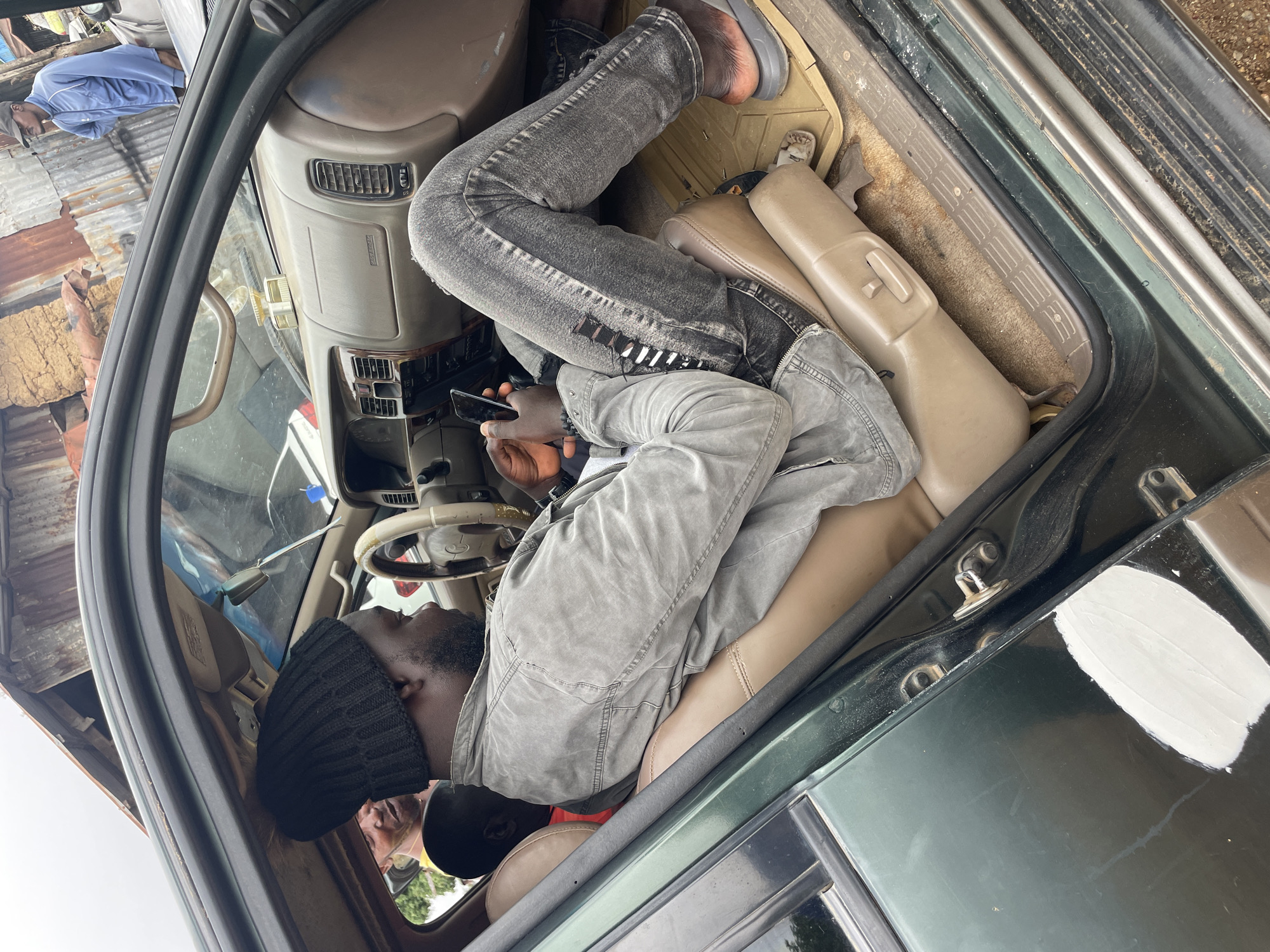 The image shows the interior of a dark green SUV in New Stadium, Nigeria. A man wearing a black beanie and a grey jacket is seated in the driver's seat, looking at his phone. His legs are extended, and he is wearing distressed grey jeans and sandals. Another person, partially visible, is in the passenger seat, wearing a yellow shirt. Outside the car, a man in a blue jacket and a baseball cap stands near a corrugated metal fence. The car's interior appears worn, with visible damage to the door panel, indicated by a patch of white filler. The environment outside suggests a somewhat rural or unkempt urban setting, with visible debris and a rustic-looking building.