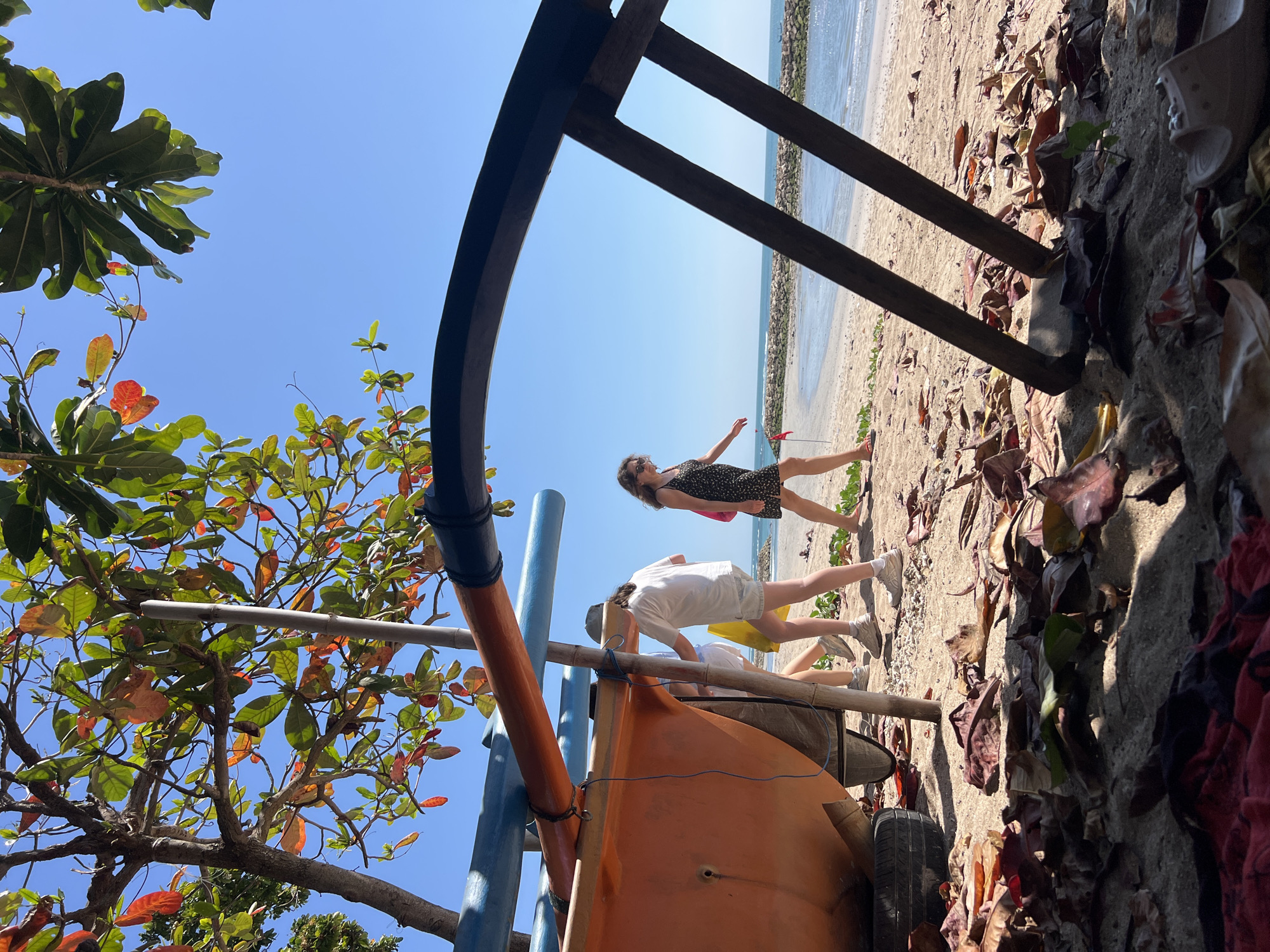 In Kuta, Indonesia, a beach scene unfolds under a clear blue sky. In the foreground, fallen leaves and sand litter the ground, with the weathered orange hull of a boat partially visible on the left. A dark, curved wooden structure frames the right side of the scene. Two individuals are walking on the sandy shore in the mid-ground. One, wearing a white t-shirt and light shorts, carries a yellow bag and walks away from the camera. To their right, a woman in a dark floral dress walks towards the camera. Beyond them, the gentle waves of the ocean meet a rocky breakwater, with a faint horizon line dividing the sea from the sky. The tropical foliage, with its vibrant green and red leaves, drapes over the scene, suggesting the lush vegetation common to the area.