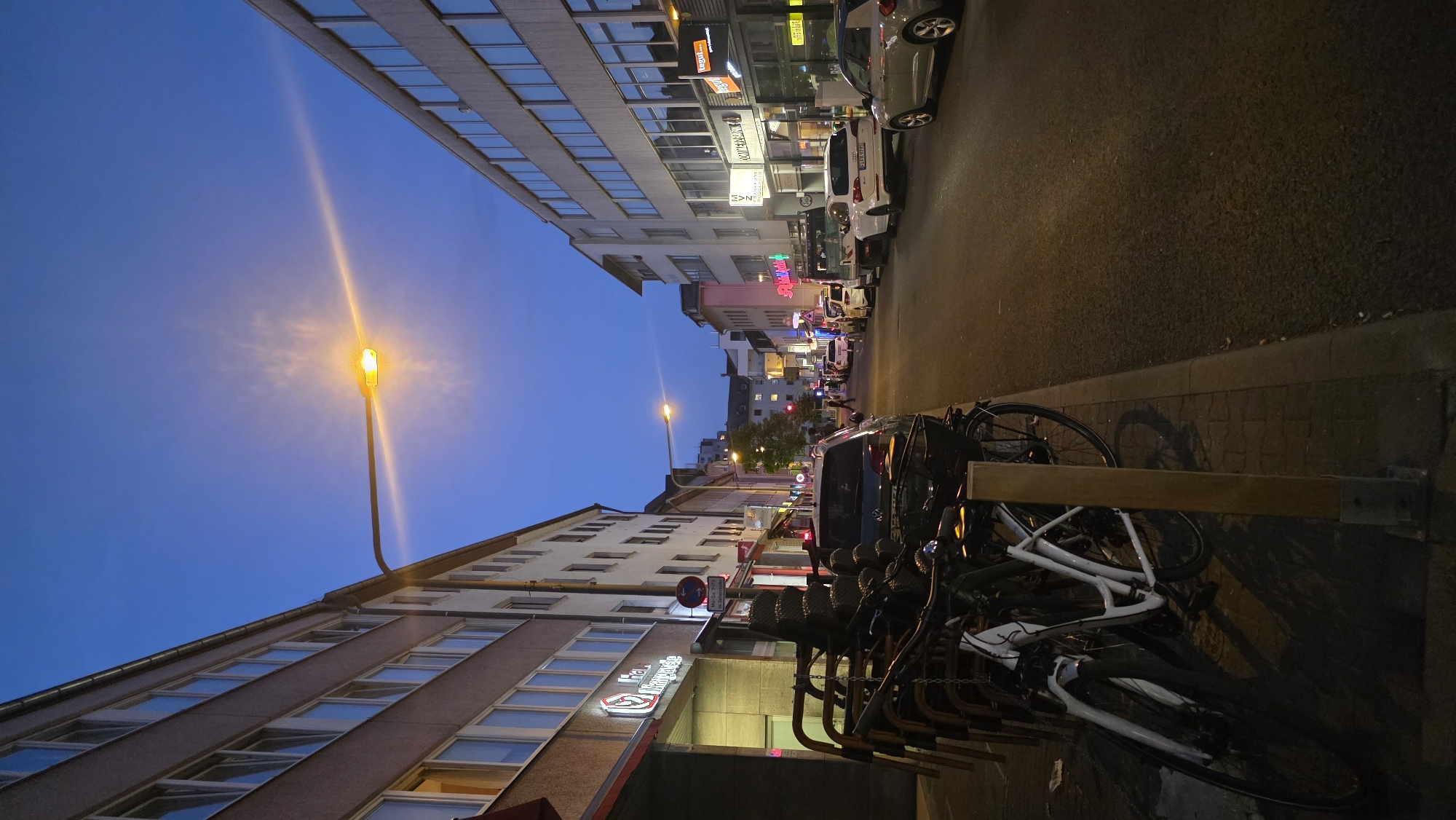 A street scene unfolds in a European city at dusk. The sky is a deep blue, transitioning from the fading daylight. A prominent street lamp casts a warm, yellow glow, its light creating a lens flare effect against the darkening sky. Buildings line both sides of the street, their facades featuring rows of windows.
On the left, a building with a light-colored facade has a commercial space on the ground floor with a sign that appears to read "Ha. Margarete." In front of this building, a stack of chairs and several bicycles are parked along the sidewalk. One white bicycle is particularly visible, leaning against a wooden post.
The street itself is paved and shows signs of traffic. Cars are parked along the right side of the road and a few are in motion. Among them are sedans and what appear to be taxis. The buildings on the right side also display commercial signage, with some illuminated signs visible, indicating businesses like "Tegui" and "MVZ Frankfurt." Pedestrians are present, walking on the sidewalks and crossing the street in the distance. The overall impression is a moderately busy urban environment at the end of the day.